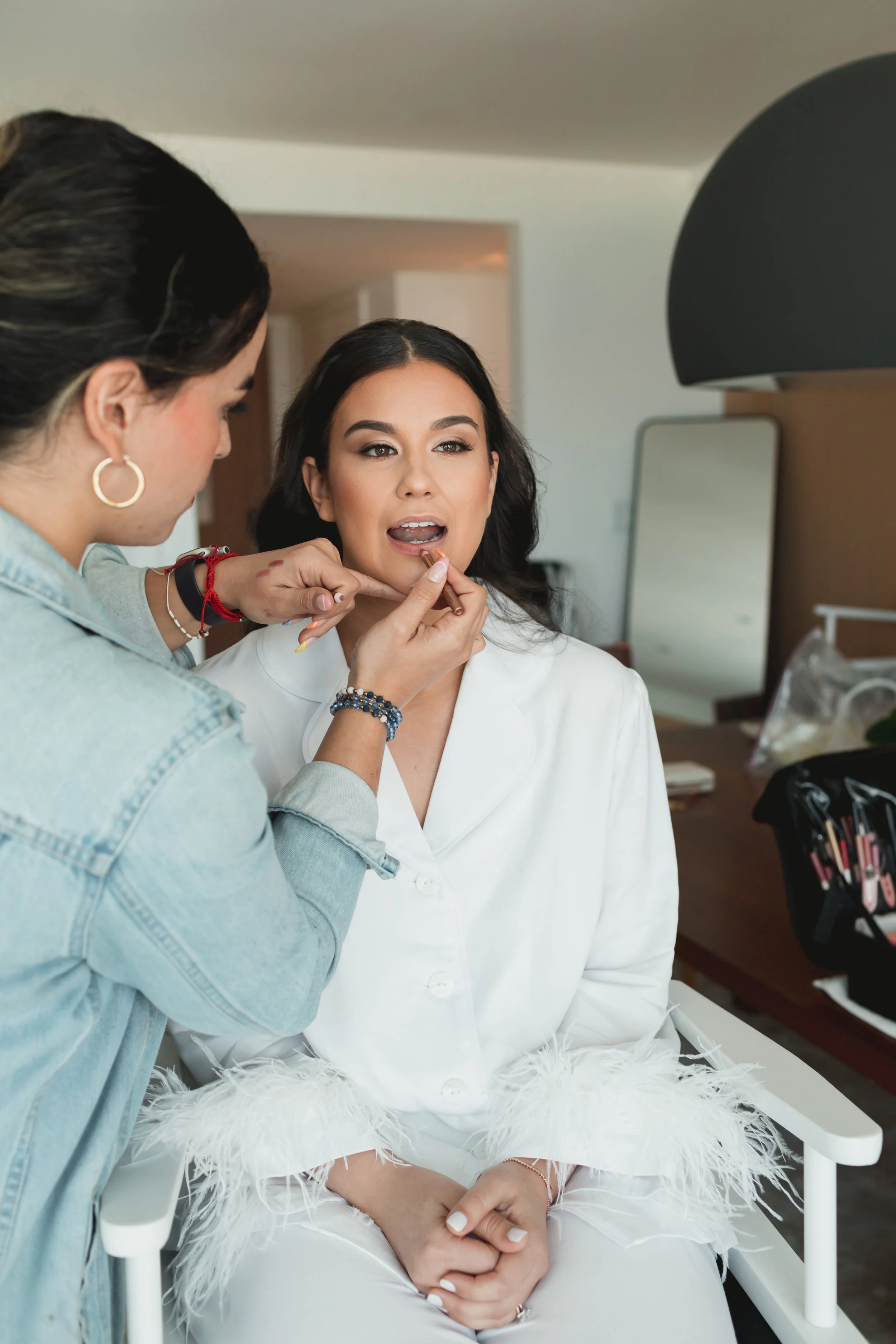 A woman in a white blazer getting her makeup done by a makeup artist in a room with a bed, a mirror, and a black lampshade.