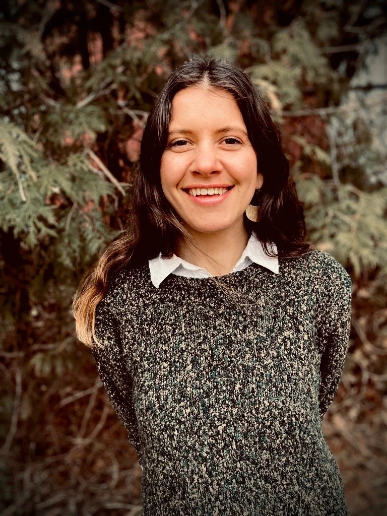 A young woman with long dark hair smiling outdoors in front of greenery, wearing a black and white textured sweater over a white collared shirt.