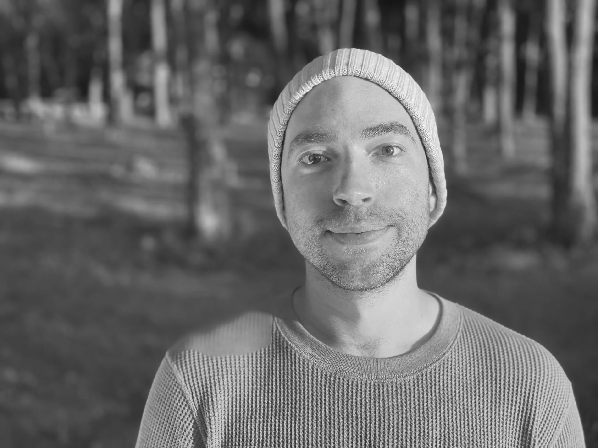A black-and-white portrait of a man wearing a knit cap, standing outdoors in a wooded area.