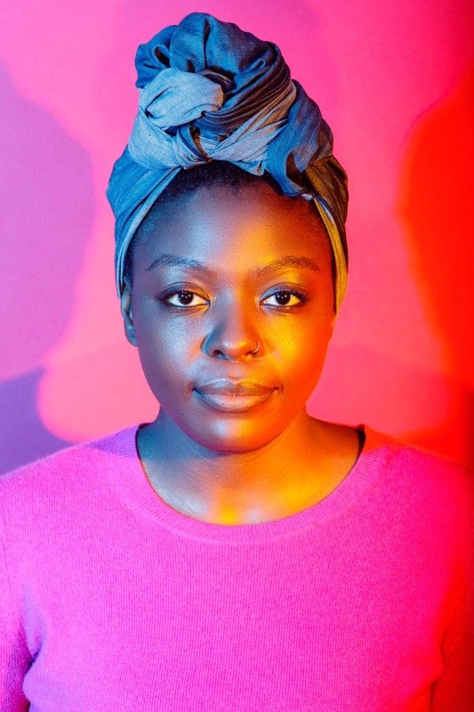 A young woman with a blue headwrap, wearing a pink sweater, is looking directly at the camera against a colorful background with pink, purple, and orange hues.