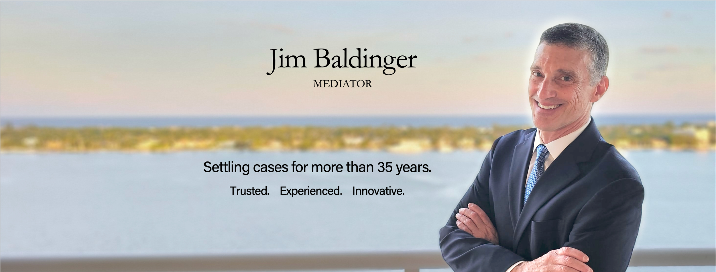 Professional headshot of Jim Baldinger, a mediator, with a blurred scenic water and landscape background, smiling in a dark suit and blue tie, overlayed by text: settling cases for more than 35 years, trusted, experienced, and innovative.