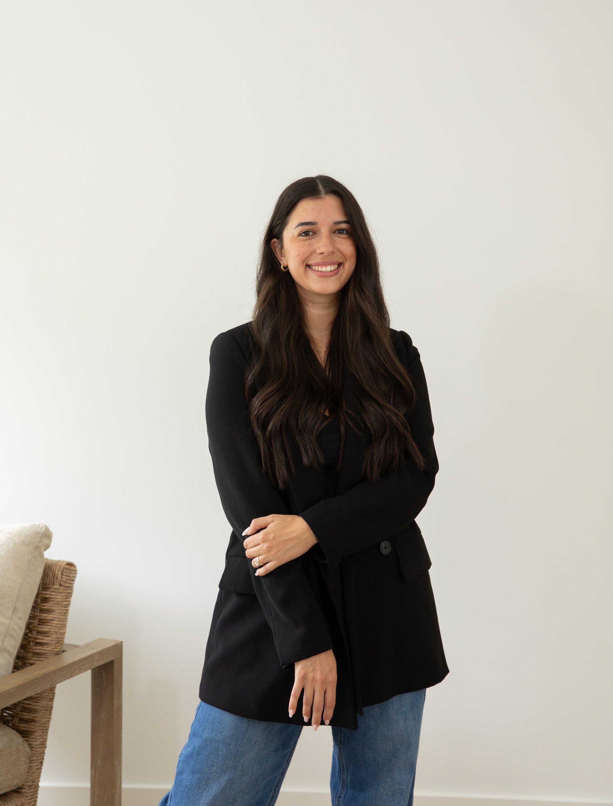 A person with long dark hair wearing a black suit stands in a modern room. There is a beige armchair with a striped cushion in the foreground and a cabinet with decorative items like a round mirror and a vase in the background.