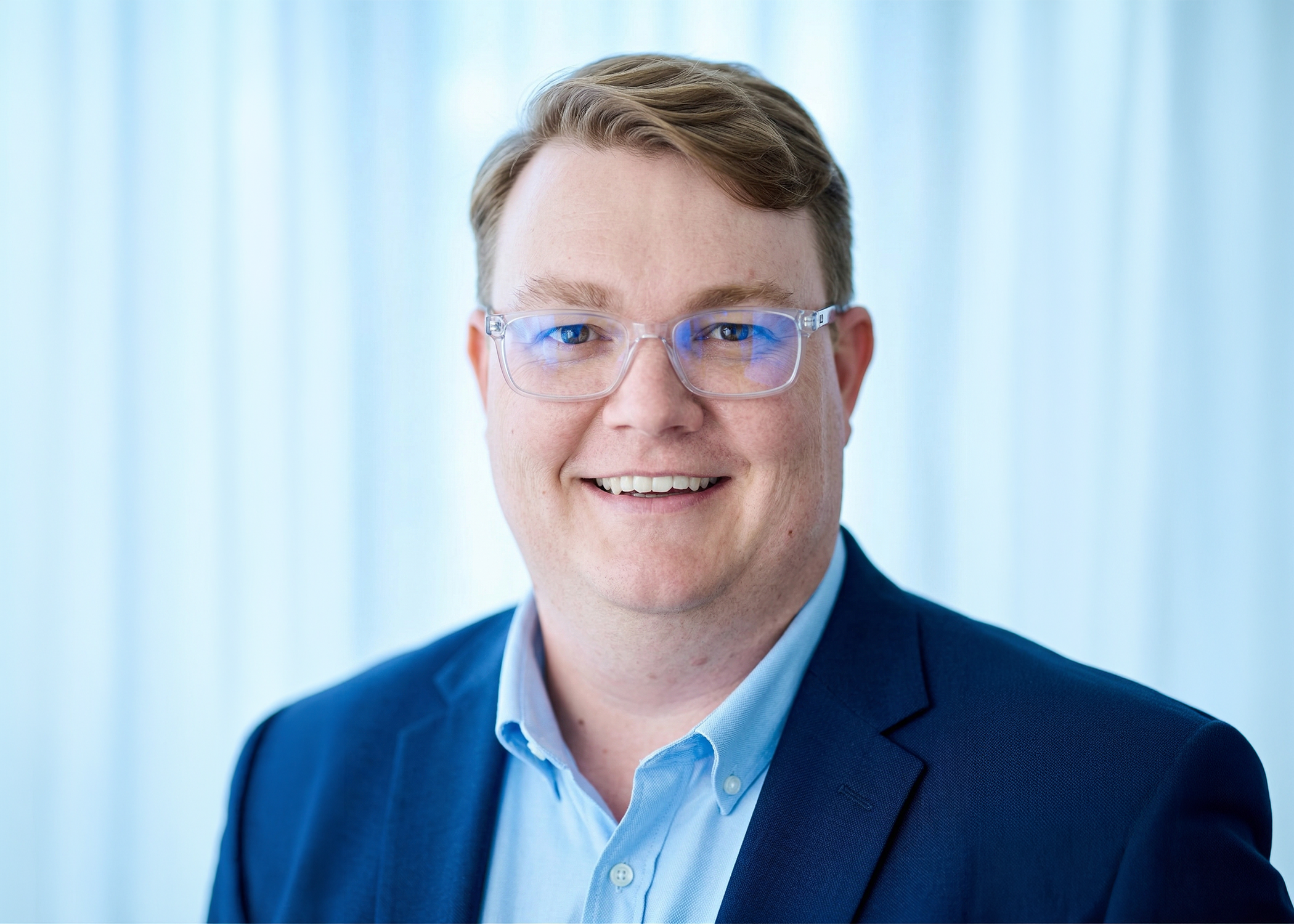 A man with glasses smiling, wearing a white shirt with a blue floral pattern, against a light blue background.