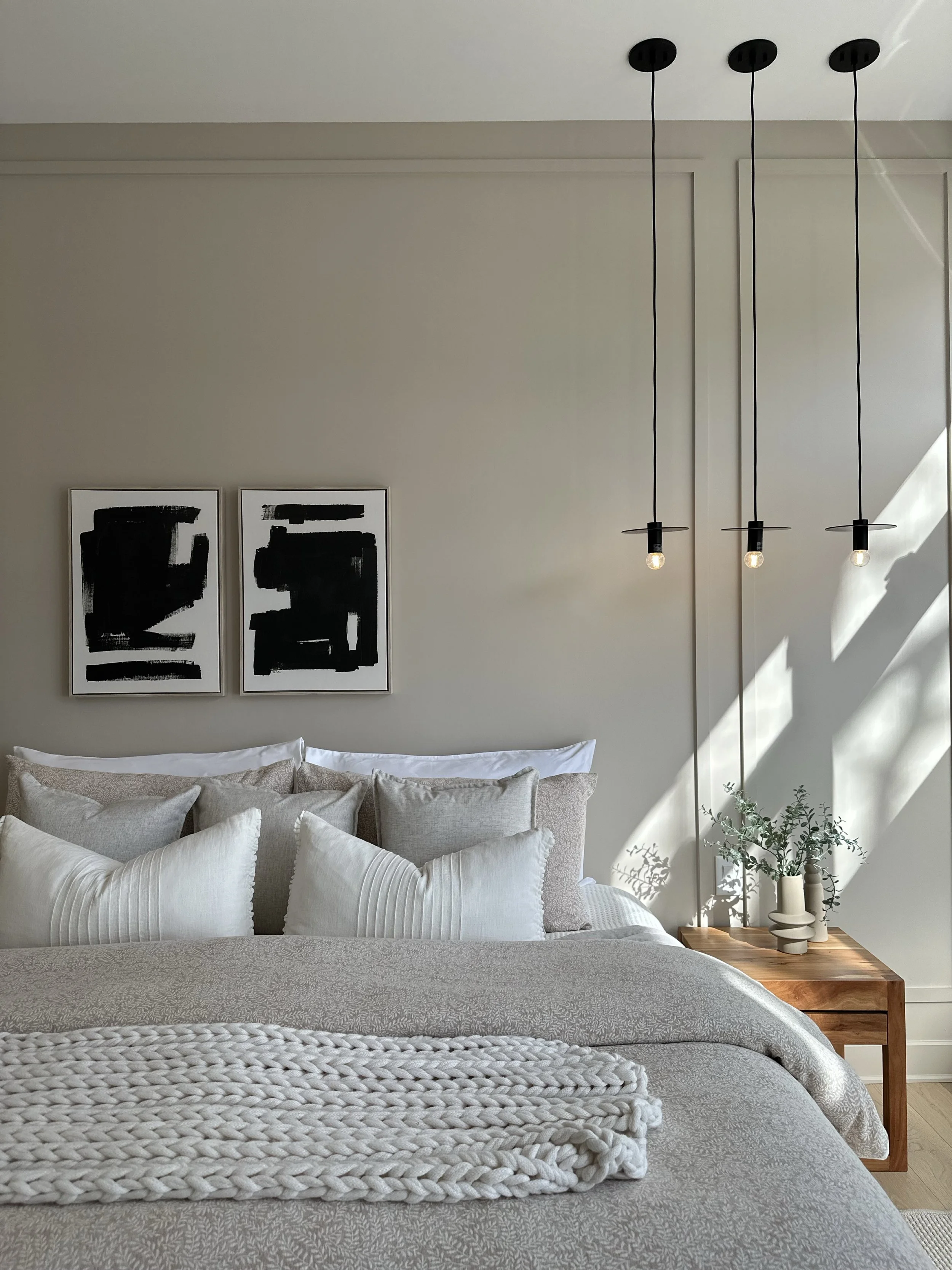 A cozy bedroom with a bed covered in light-colored bedding and multiple white pillows. On the wall above the bed, there are two black and white abstract art framed pictures. To the right, a wooden nightstand holds a collection of white vases with greenery. Three black pendant lights with exposed bulbs hang from the ceiling.