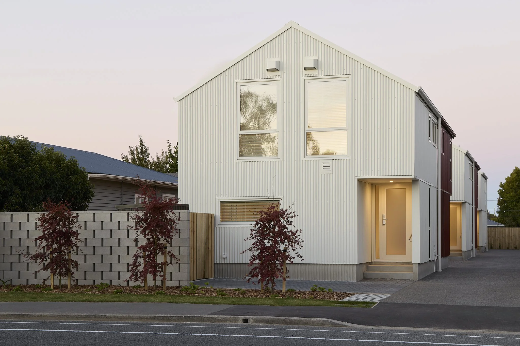 Modern two-story architectual townhouse development with vertical corrugate cladding and matching roof, enclosed backyard with garden, and neighboring homes on a sunny day.