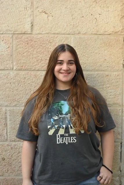A young woman with long brown hair smiling, standing in front of a beige stone wall, wearing a black The Beatles T-shirt.