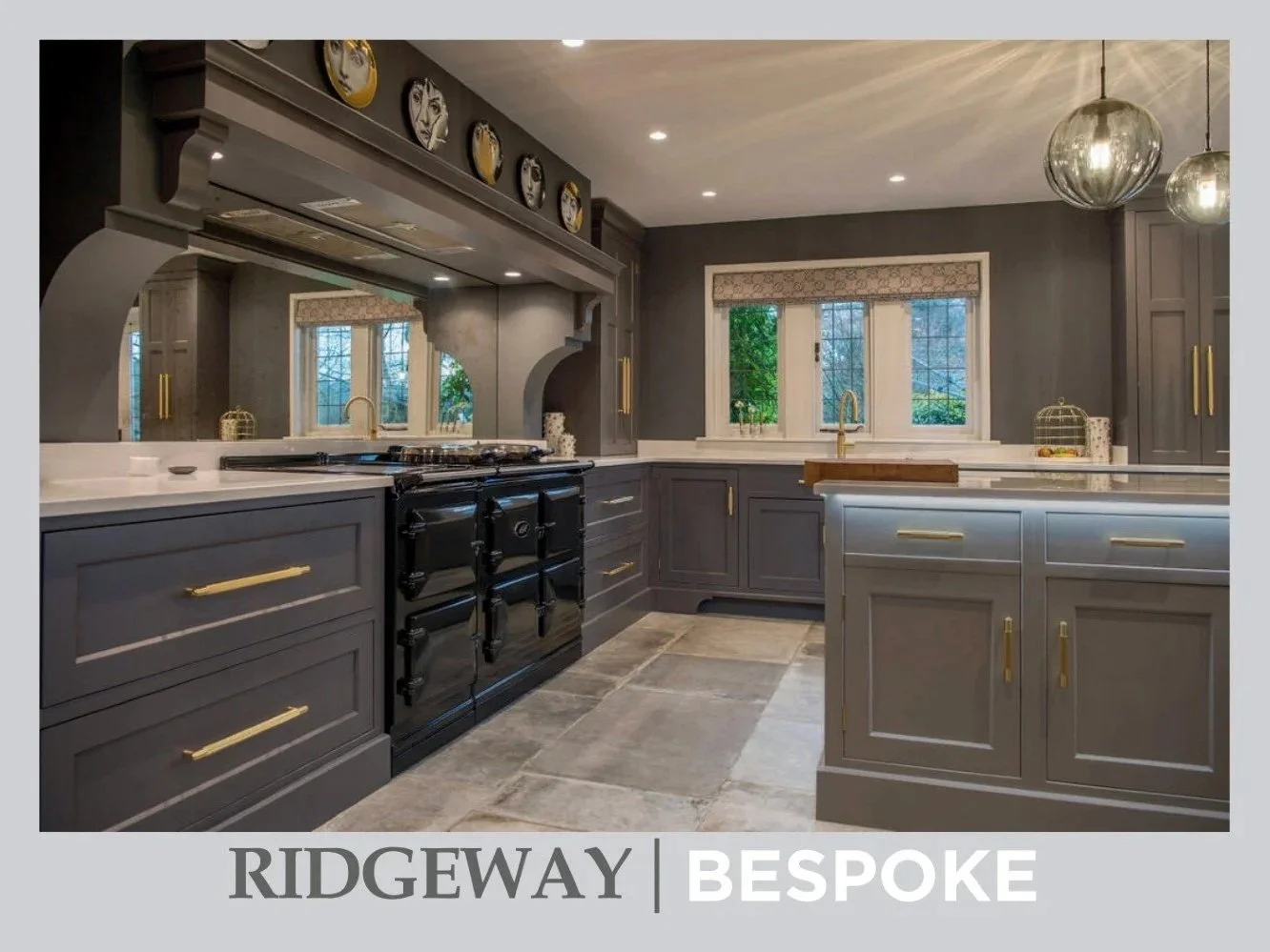 Interior view of a traditional kitchen with grey cabinets, a spacious island, pendant lights hanging from the ceiling, and a windows with greenery outside.