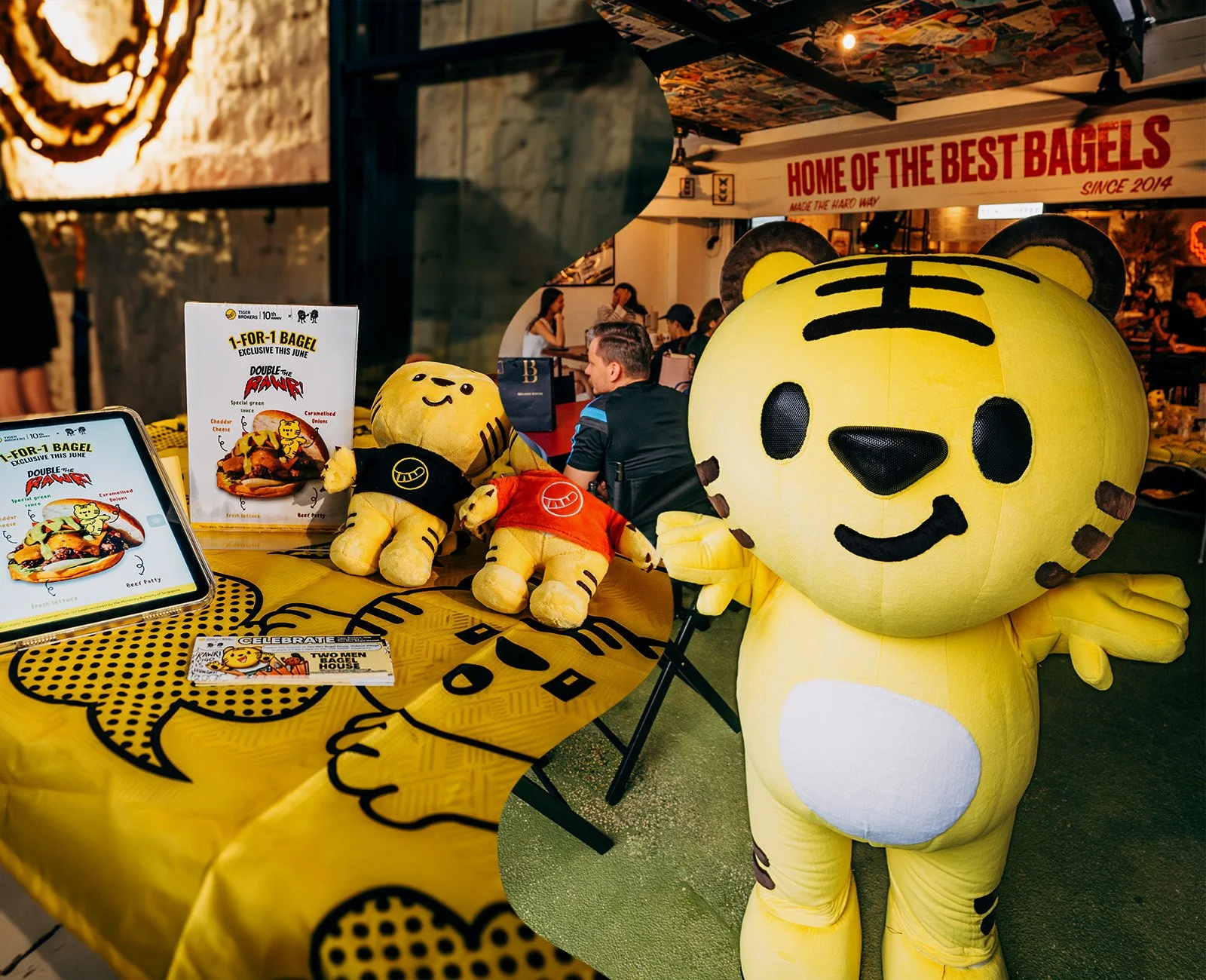 A yellow tiger mascot costume standing next to a table with plush toys and promotional signage in a restaurant or cafe setting.