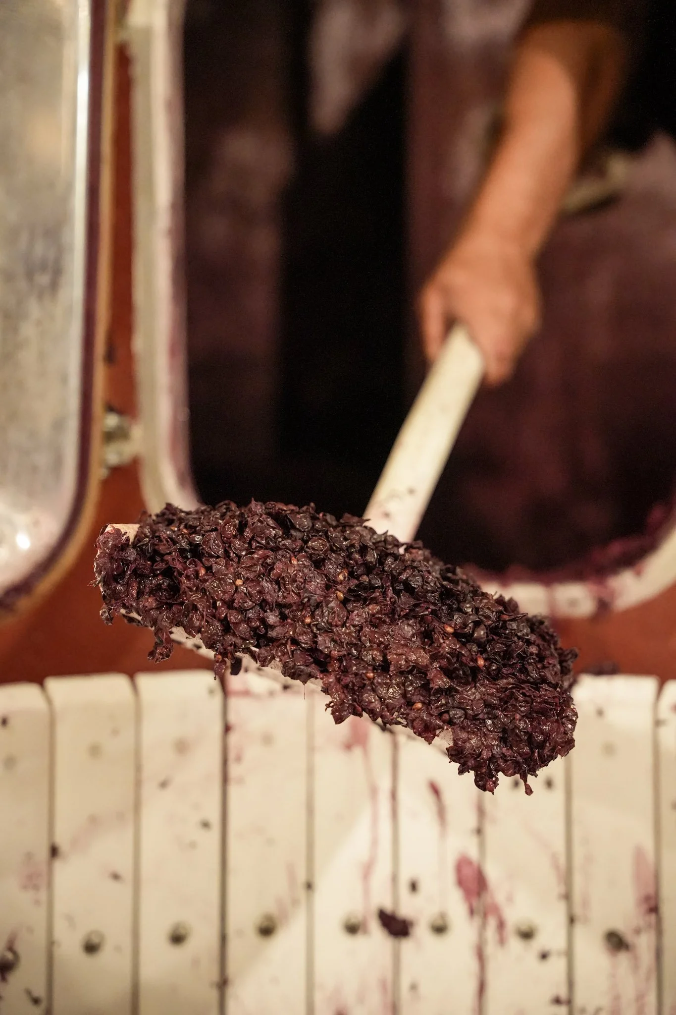 Une cuillère remplie de mûres noires ou de cassis, avec une personne en arrière-plan utilisant un ustensile pour écraser des fruits dans un récipient.