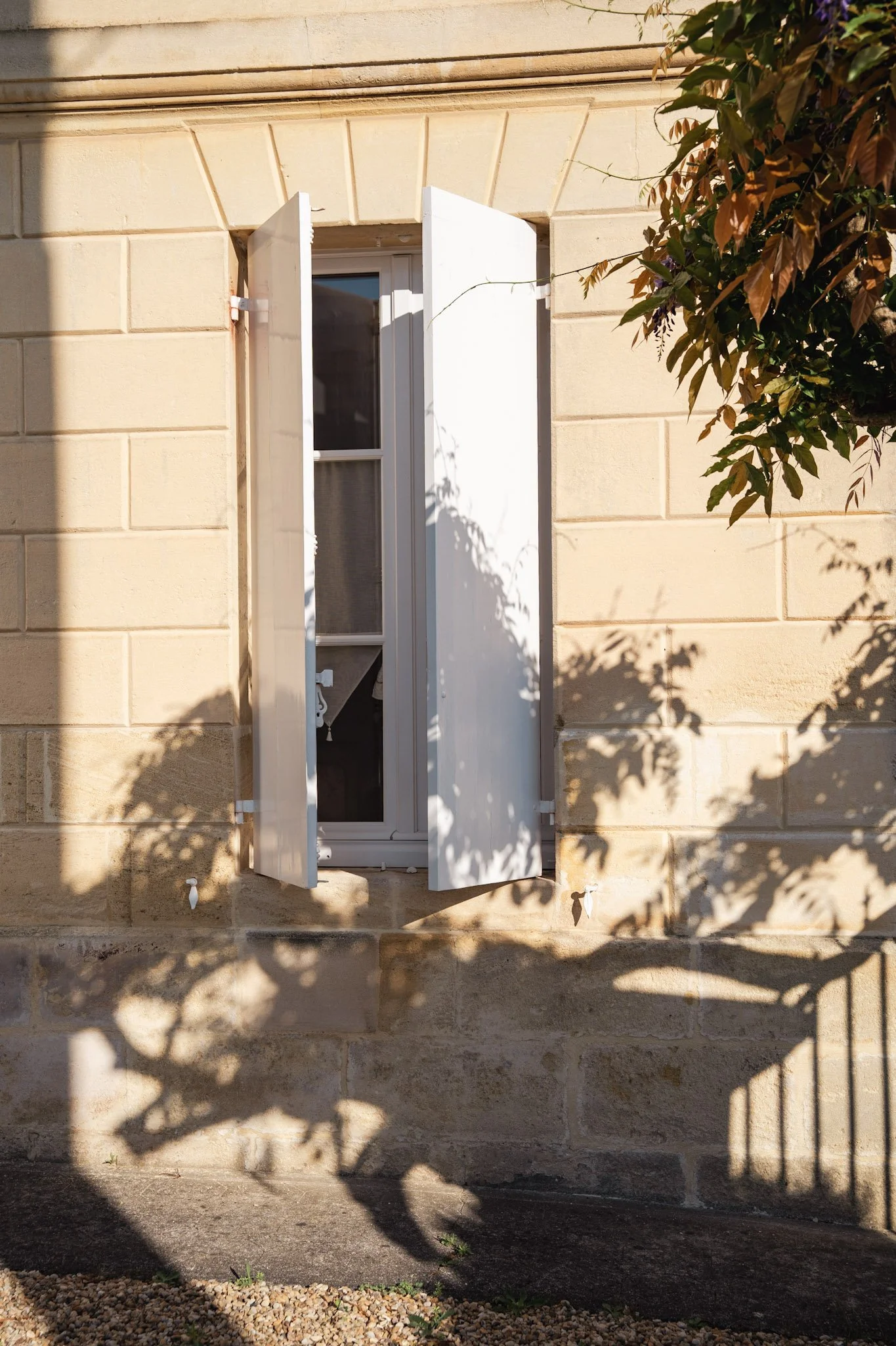 Fenêtre avec volets blancs dans un mur en pierre beige, avec des ombres de feuilles d'arbre projetées au sol.