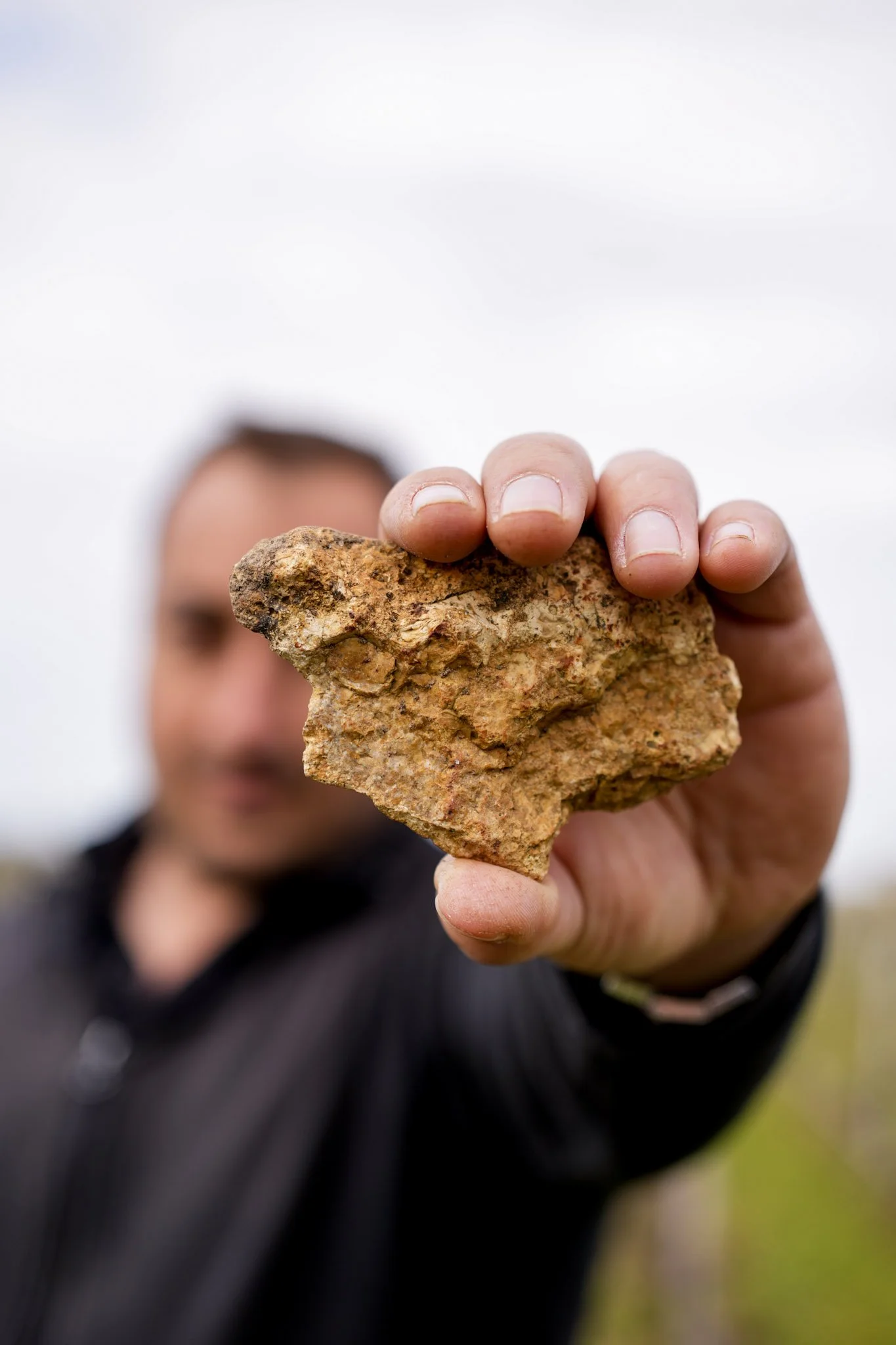 Une personne tient un gros morceau de roche ou de minerai devant l'appareil, floue en arrière-plan avec le ciel nuageux.