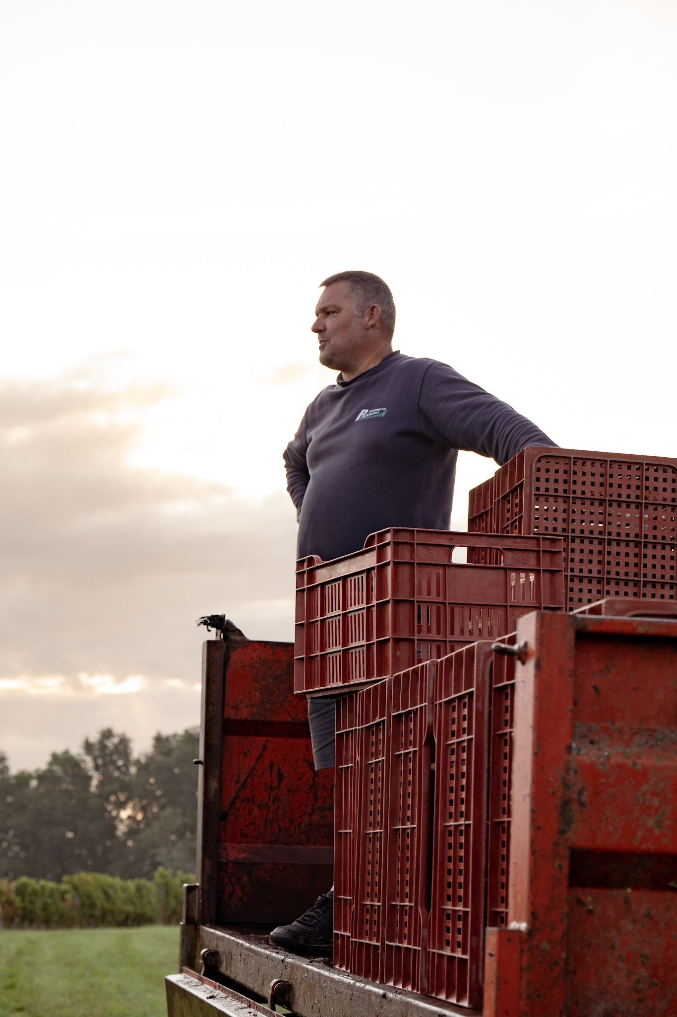 Homme sur un véhicule de ferme, entouré de caisses en plastique rouges, à l'extérieur avec un ciel nuageux en arrière-plan.