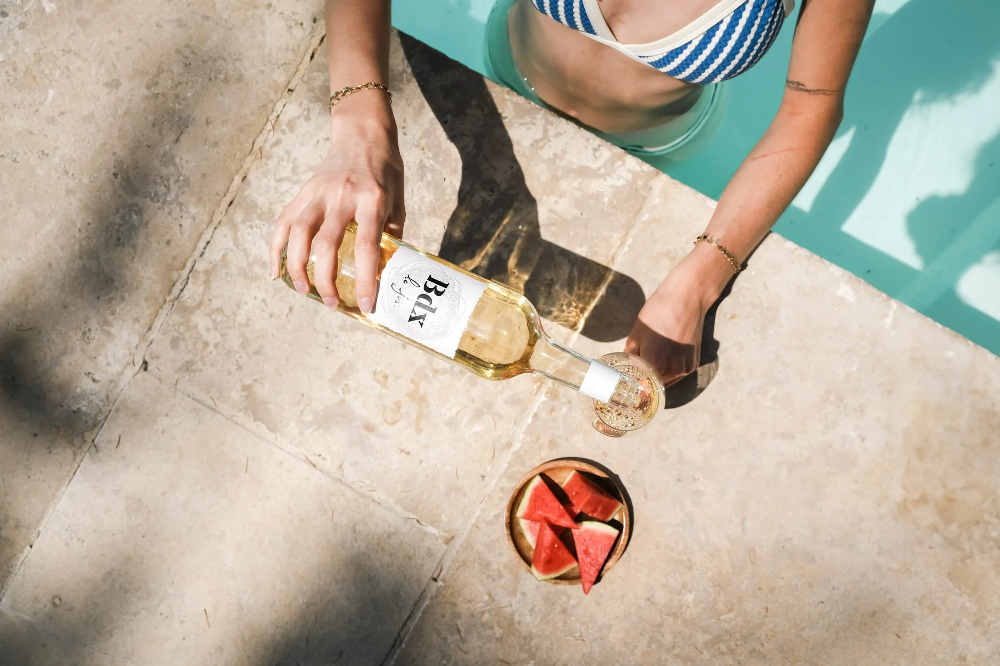 Une personne tenant une bouteille de vin blanc en sirotant au bord d'une piscine, avec un bras doré, portant un maillot de bain à rayures bleues et blanches et un bracelet.