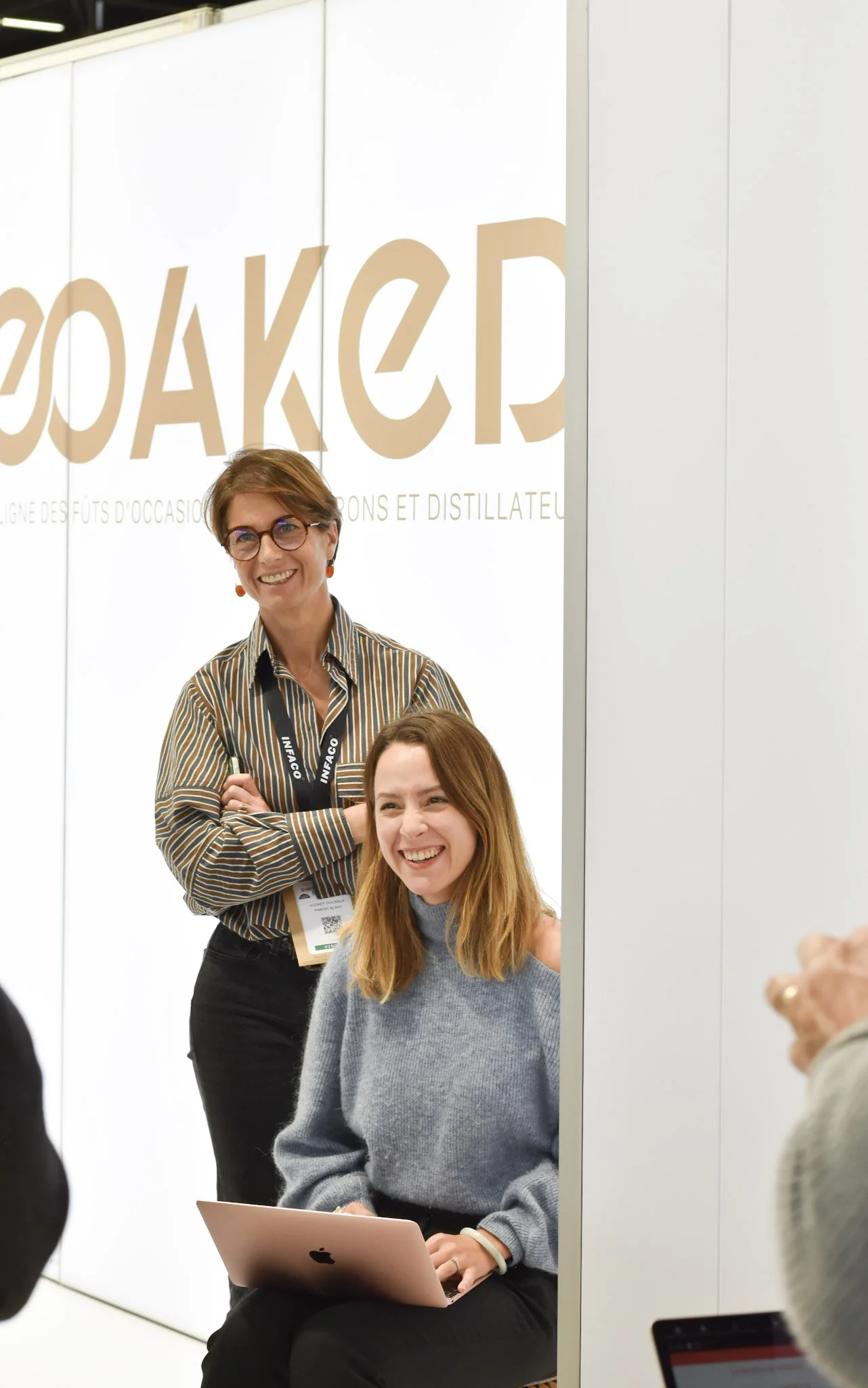 Deux femmes en réunion, l'une assise avec un ordinateur portable et l'autre debout, toutes deux souriantes, dans un espace professionnel avec un panneau blanc avec du texte en arrière-plan.