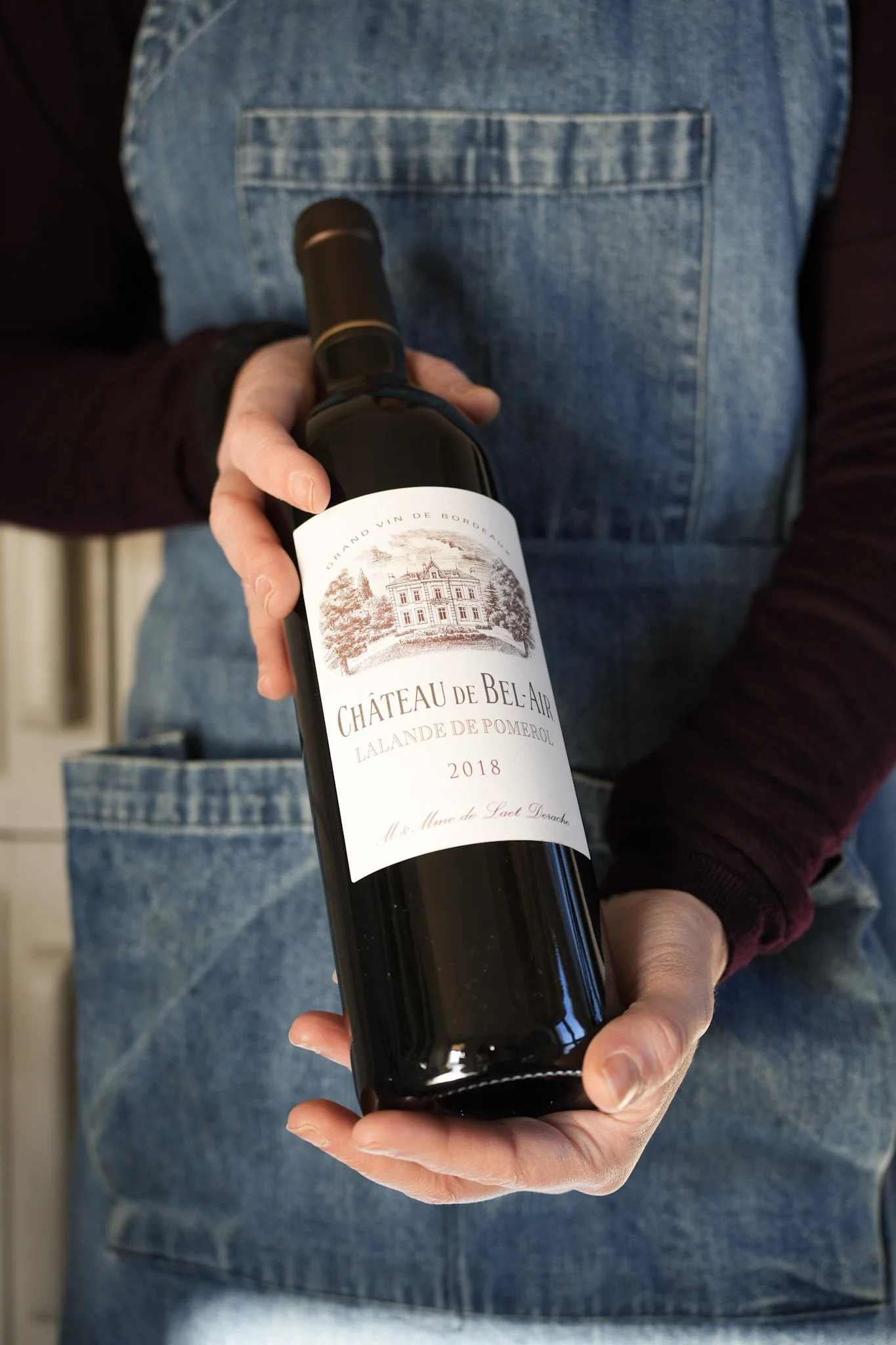 Personne tenant une bouteille de vin rouge Château de Bel-Air, année 2018, en portant un tablier en denim.