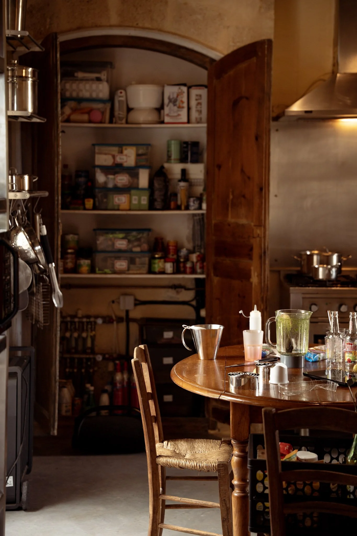 Une cuisine rustique avec un table en bois, des chaises, un étagère ouverte avec des boîtes de nourriture, et divers ustensiles de cuisine.