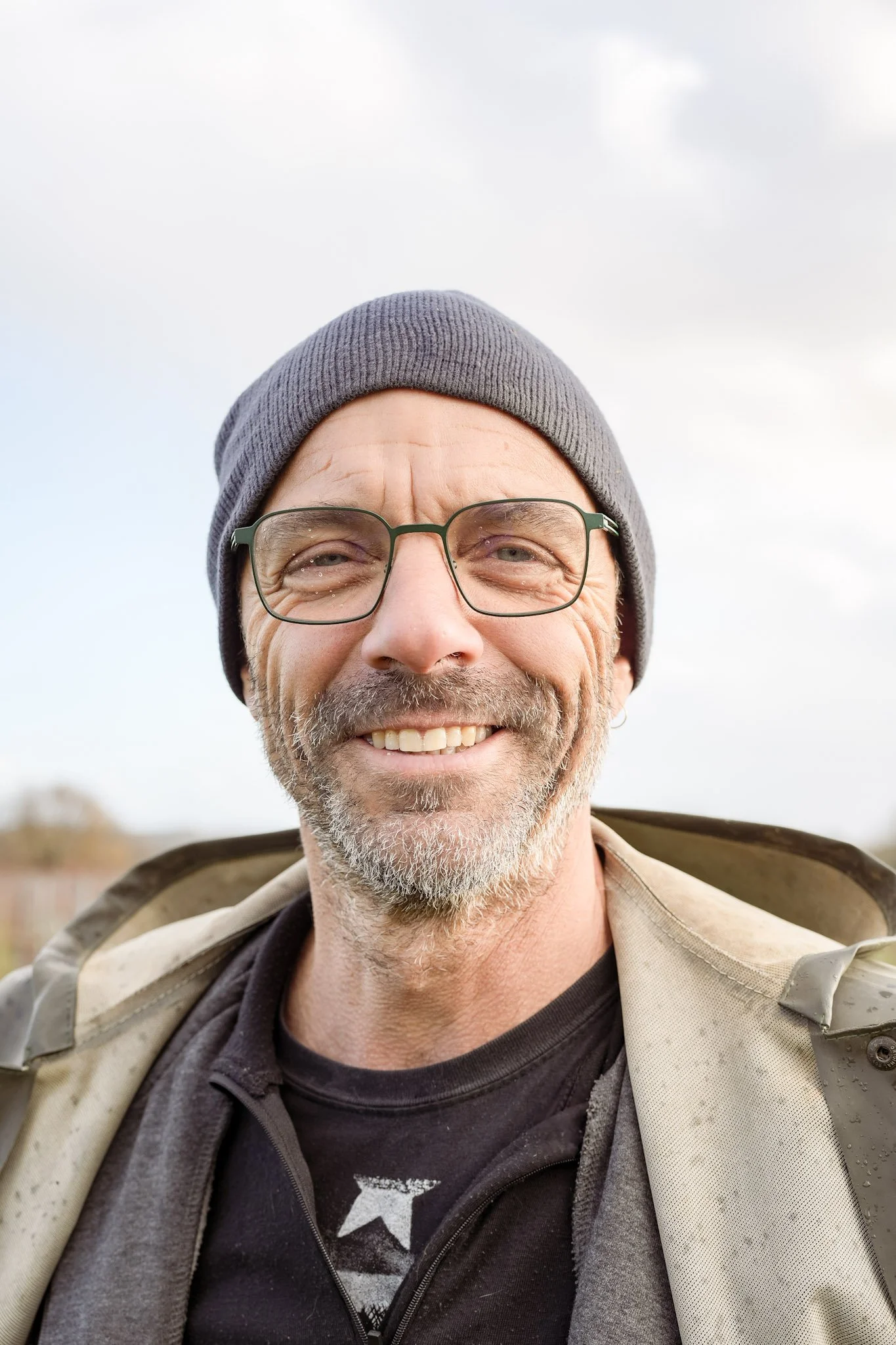 Homme souriant avec lunettes, bonnet gris, barbe grise, portant un manteau beige et un t-shirt noir, en plein air.