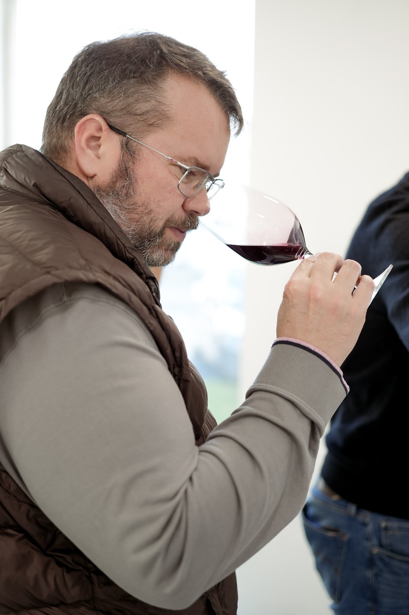 Un homme avec des lunettes, une barbe grise, portant un haut marron et un sweat gris, sentant un verre de vin rouge."