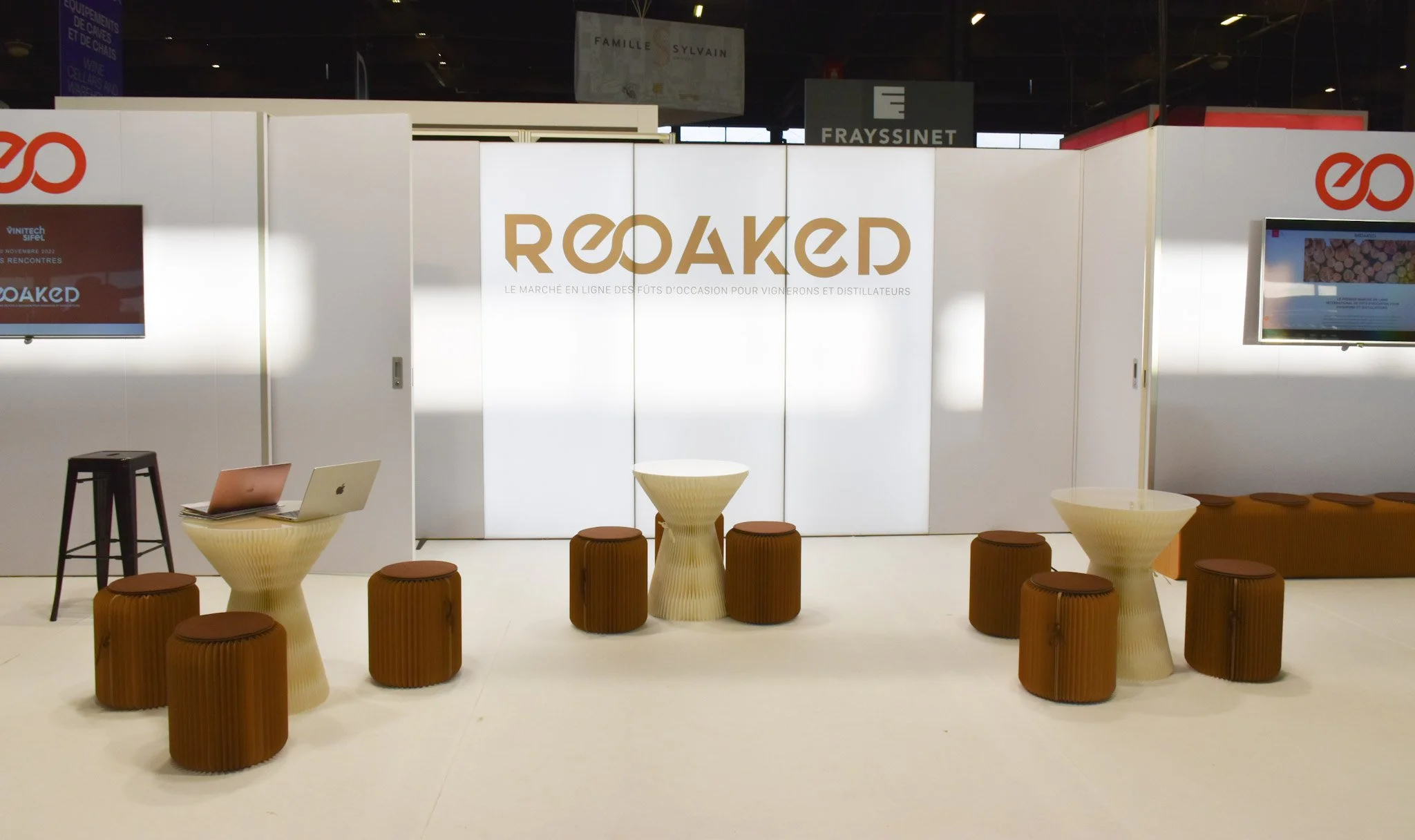 Stand d'exposition avec logo 'Roaked' et mobilier en bois, deux télévisions et deux tables avec ordinateurs portables.