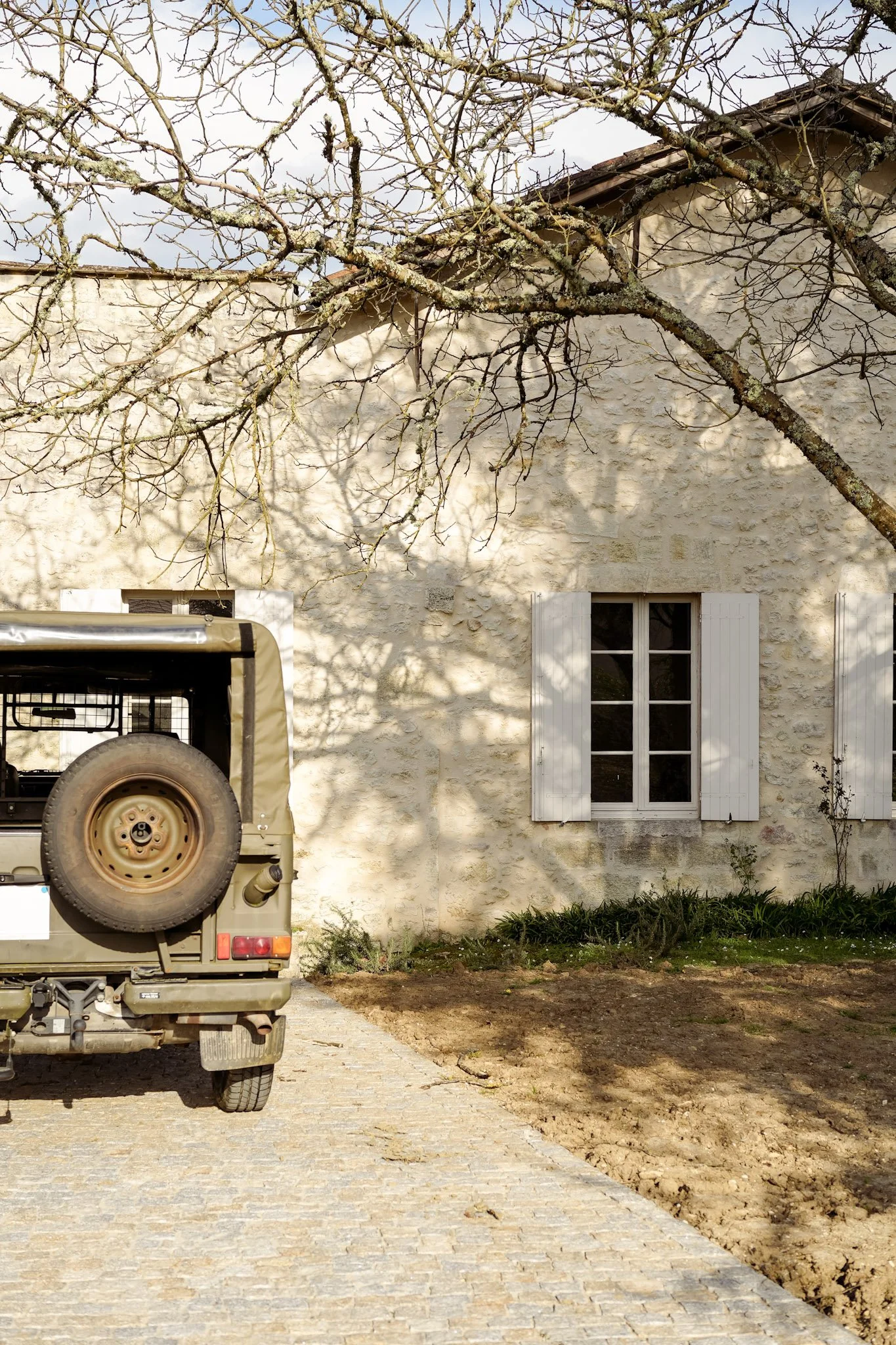 Une maison en pierre avec des volets blancs, un arbre sans feuilles projetant des ombres sur le mur, et une voiture garée sur un chemin en pavés.
