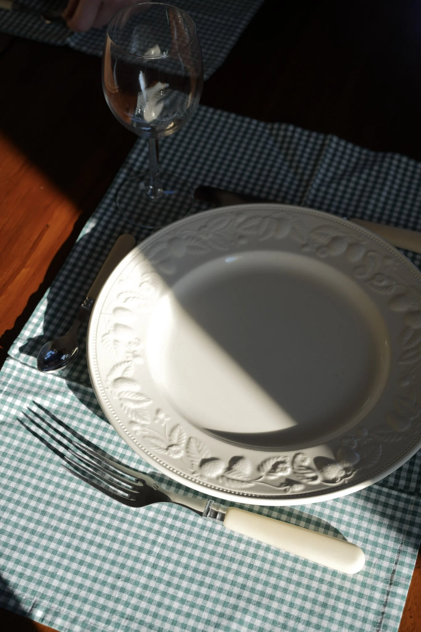 Une table dressée avec une assiette blanche en faïence avec un motif de feuilles, une fourchette avec manche en ivoire, une cuillère en métal, un verre à vin transparent, et une nappe à carreaux verts et blancs, avec un éclairage naturel créant une ombre sur la table.
