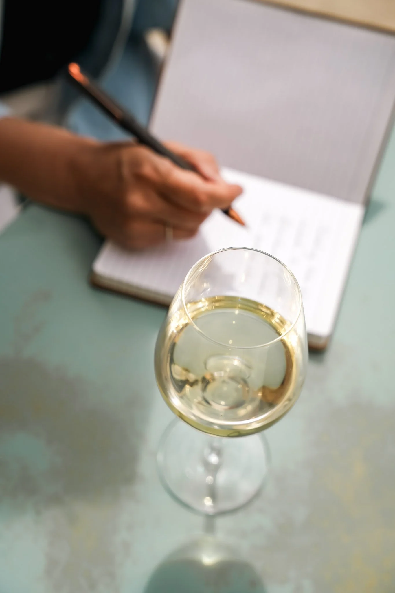Verre de vin blanc sur une table, avec une personne en arrière-plan prenant des notes dans un carnet.