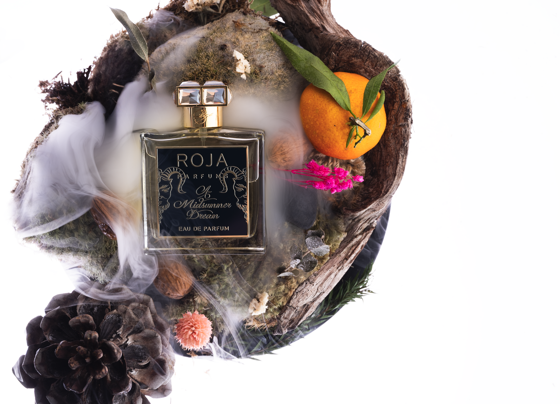 Ein Parfümflasche mit dem Label 'ROJA Parfums & Midsummer Dream', umgeben von Moos, einem orangefarbenen und pinken Blumen, Tannenzapfen, einem Stück Holz, gefrorenen Beeren, Eukalyptusblättern und einer glühenden Kerze, auf einem hölzernen Herzform-