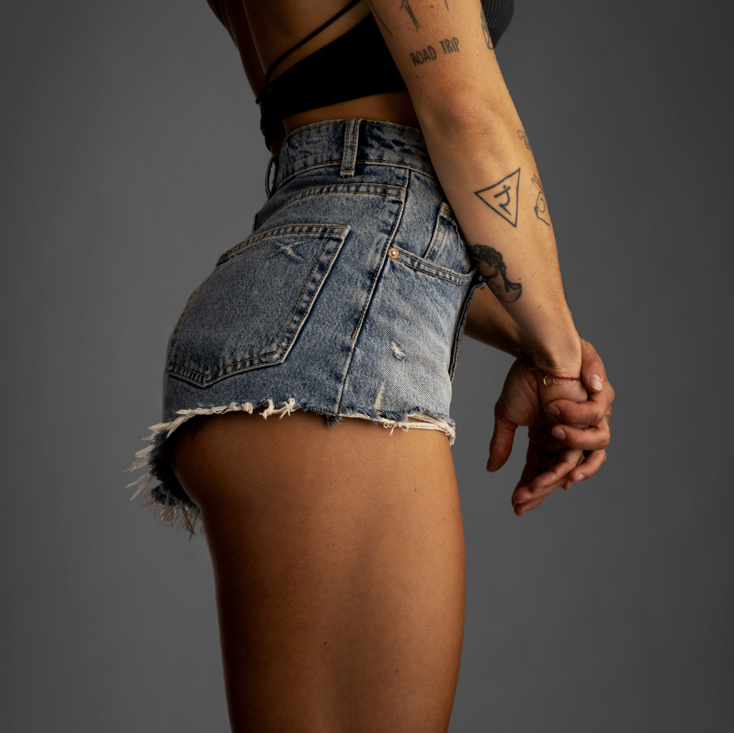 Nahaufnahme des seitlichen Teils eines weiblichen Körpers, das eine blaue Denim-Shorts und einen schwarzen Crop-Top trägt, mit sichtbaren Tattoos am Arm und einer Hand, die nach hinten zeigt, vor einem grauen Hintergrund.