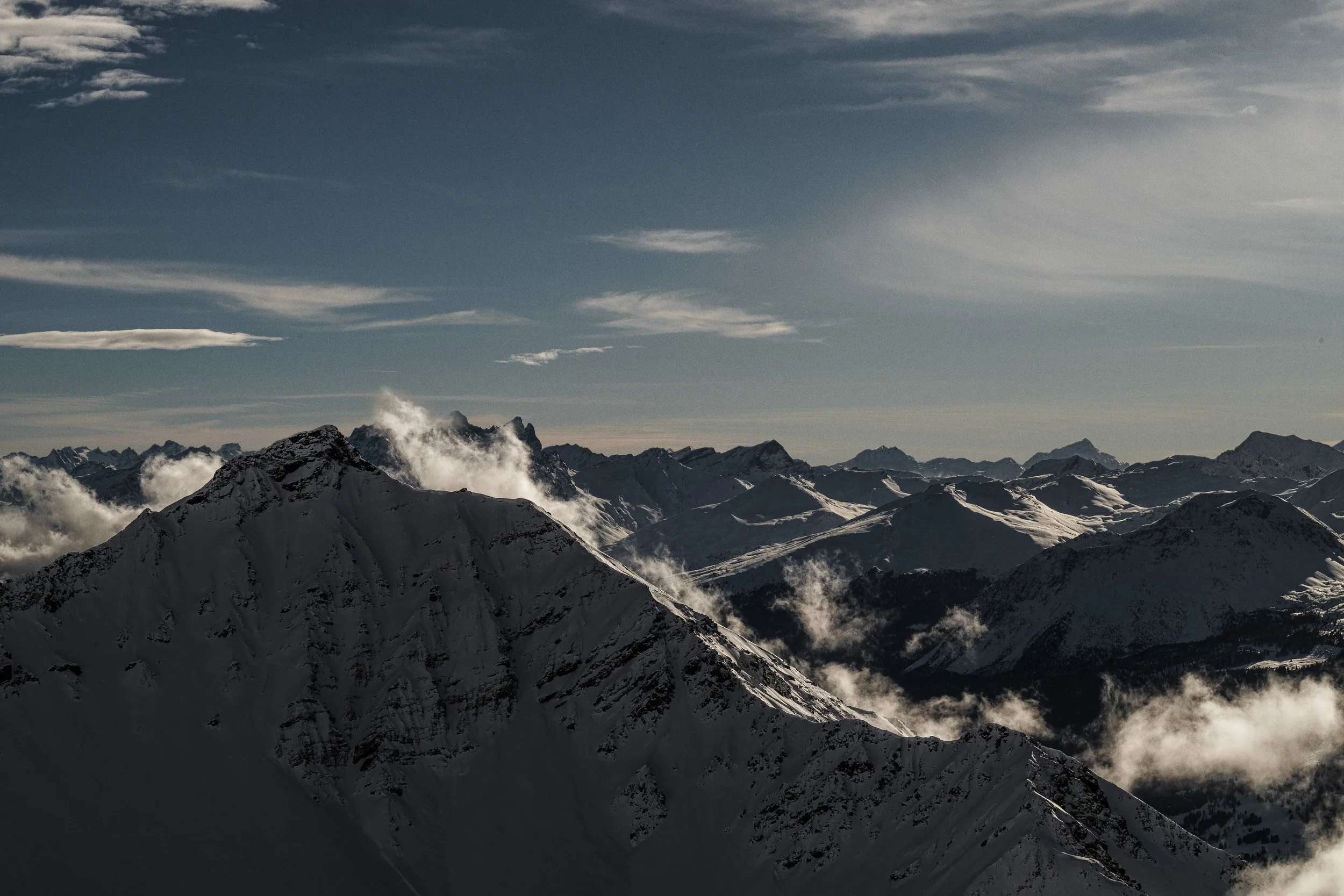 Bergkette mit schneebedeckten Gipfeln bei Sonnenaufgang oder -untergang, einige Wolken um die Berge