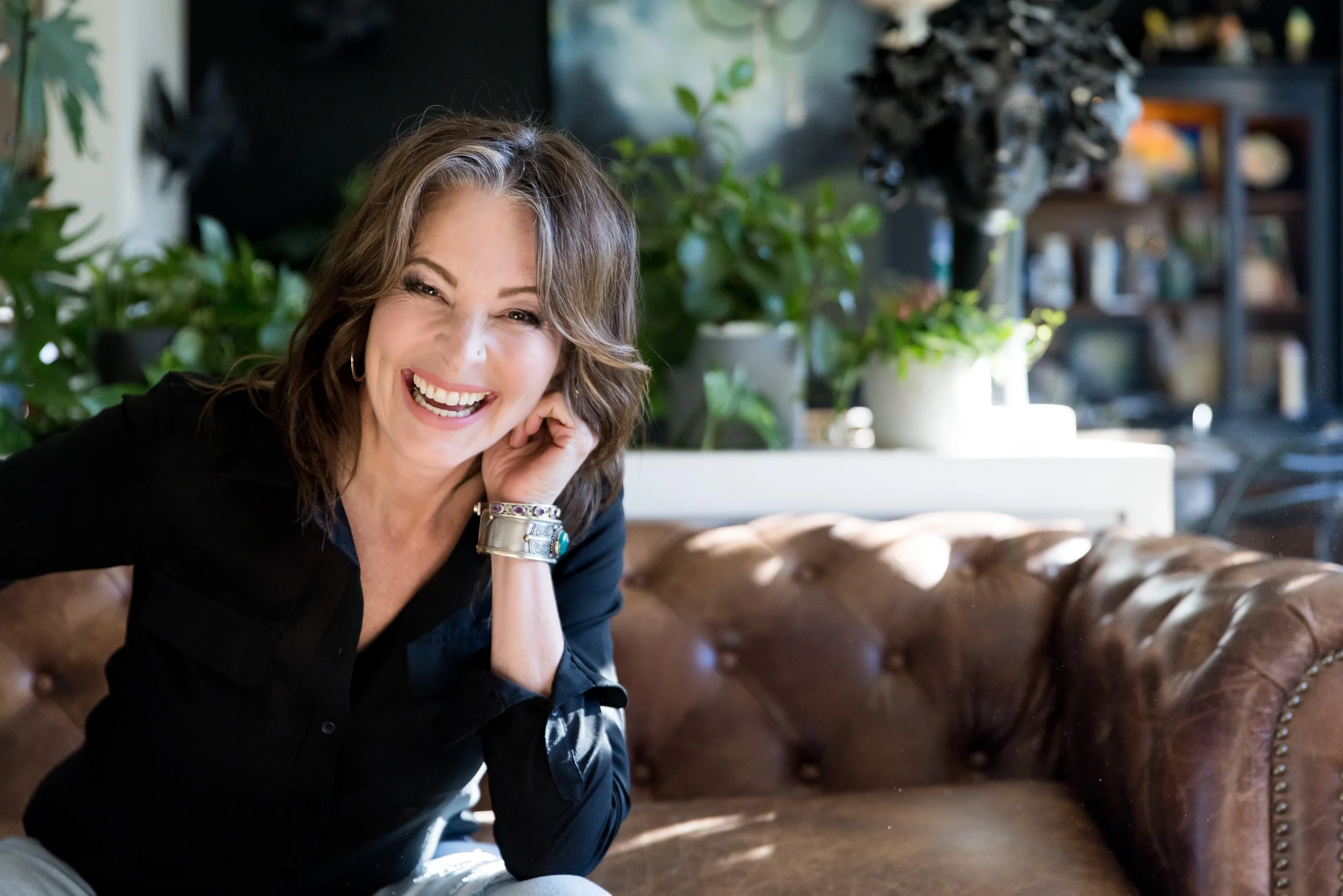 Smiling woman sitting on a brown leather couch in a room with plants and decor.
