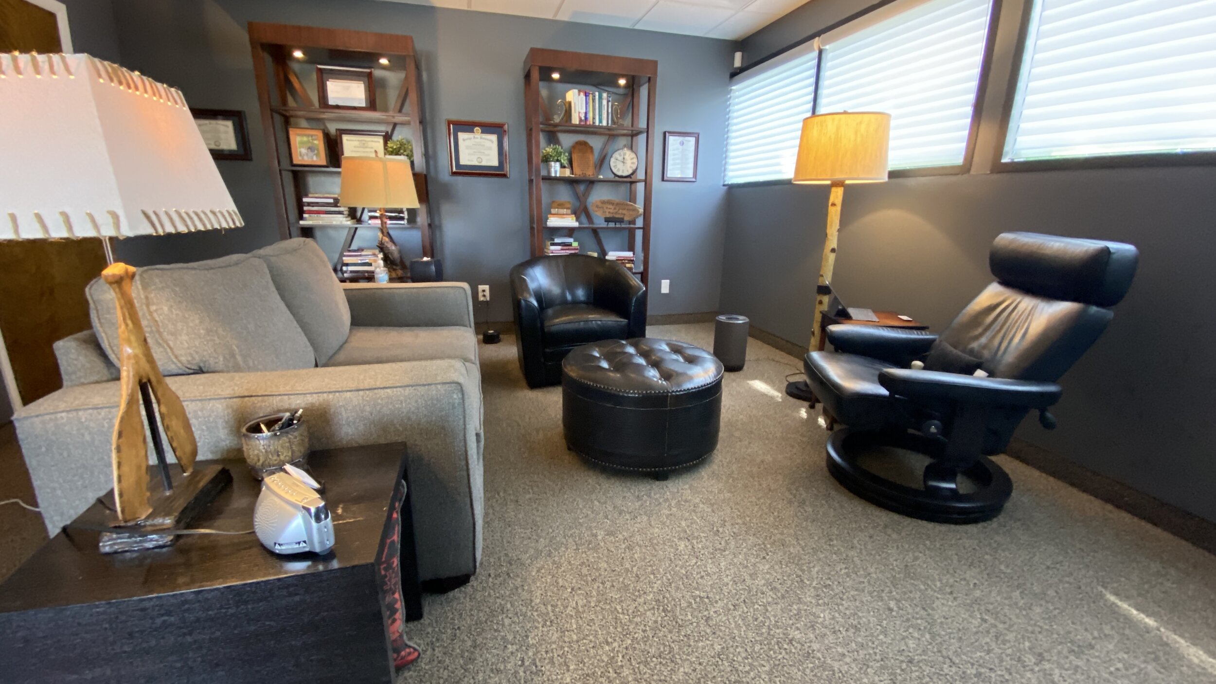 Interior of Love and Respect Counseling office with gray couch, black leather chair, round ottoman, wooden bookshelves, table lamps, and window blinds.
