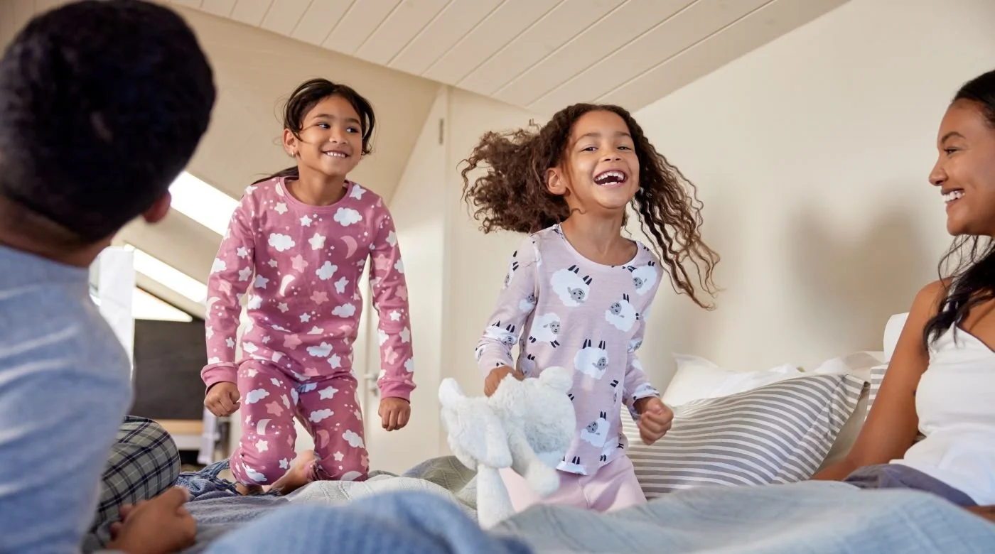 Happy Afro-American couple lying in the bed in the morning and looking at their two full of energy little girls in pygamas jumping on their parents bed