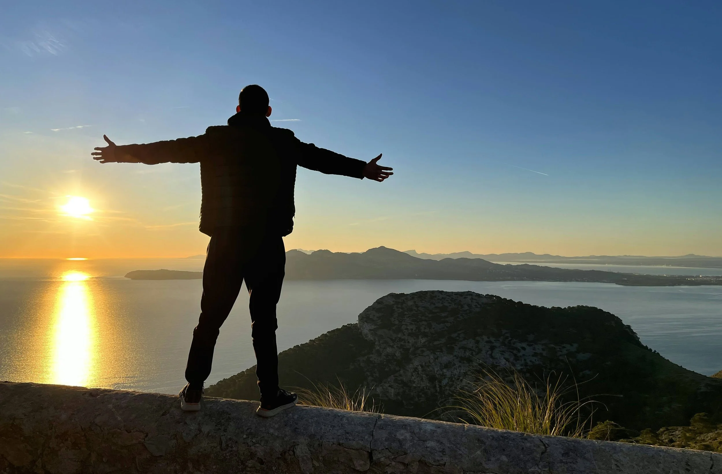 Man stands with arms outstretched on stone wall overlooking water with small mountainous islands at sunrise representing mindfulness to ease into vacation to avoid feeling guilty for taking time off work
