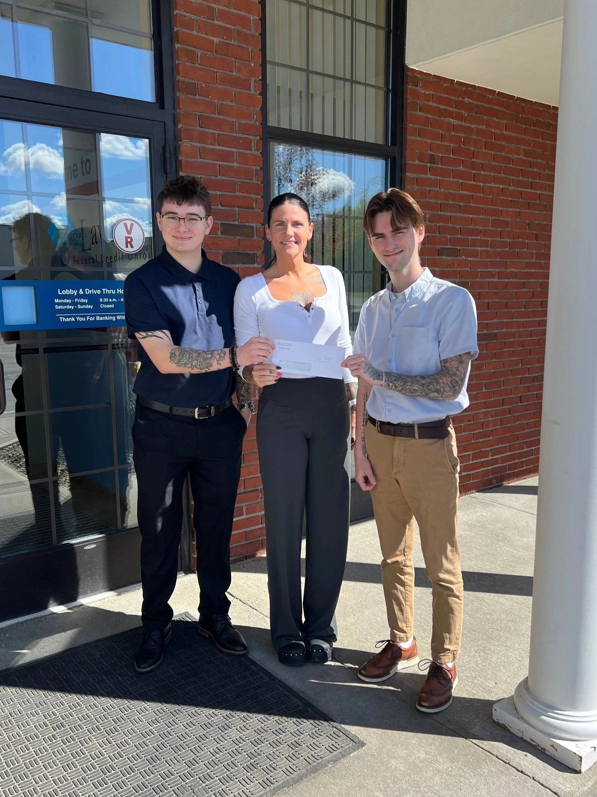 Two young adult males presenting a donation check to an adult female outside of the credit union doors.