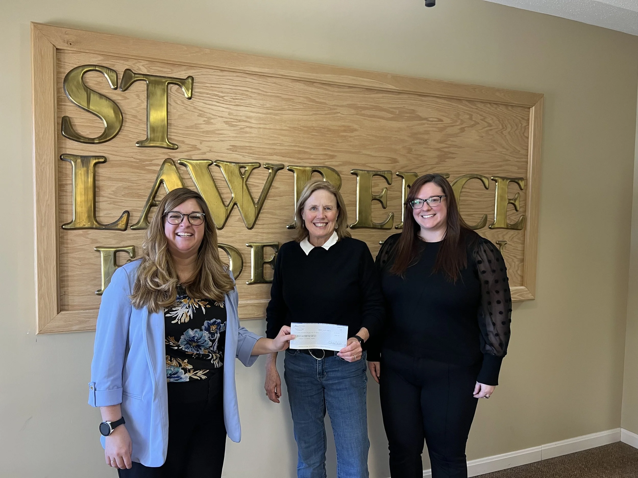 Two adult females presenting a donation check to one adult female for the Hammond Scottish Festival.