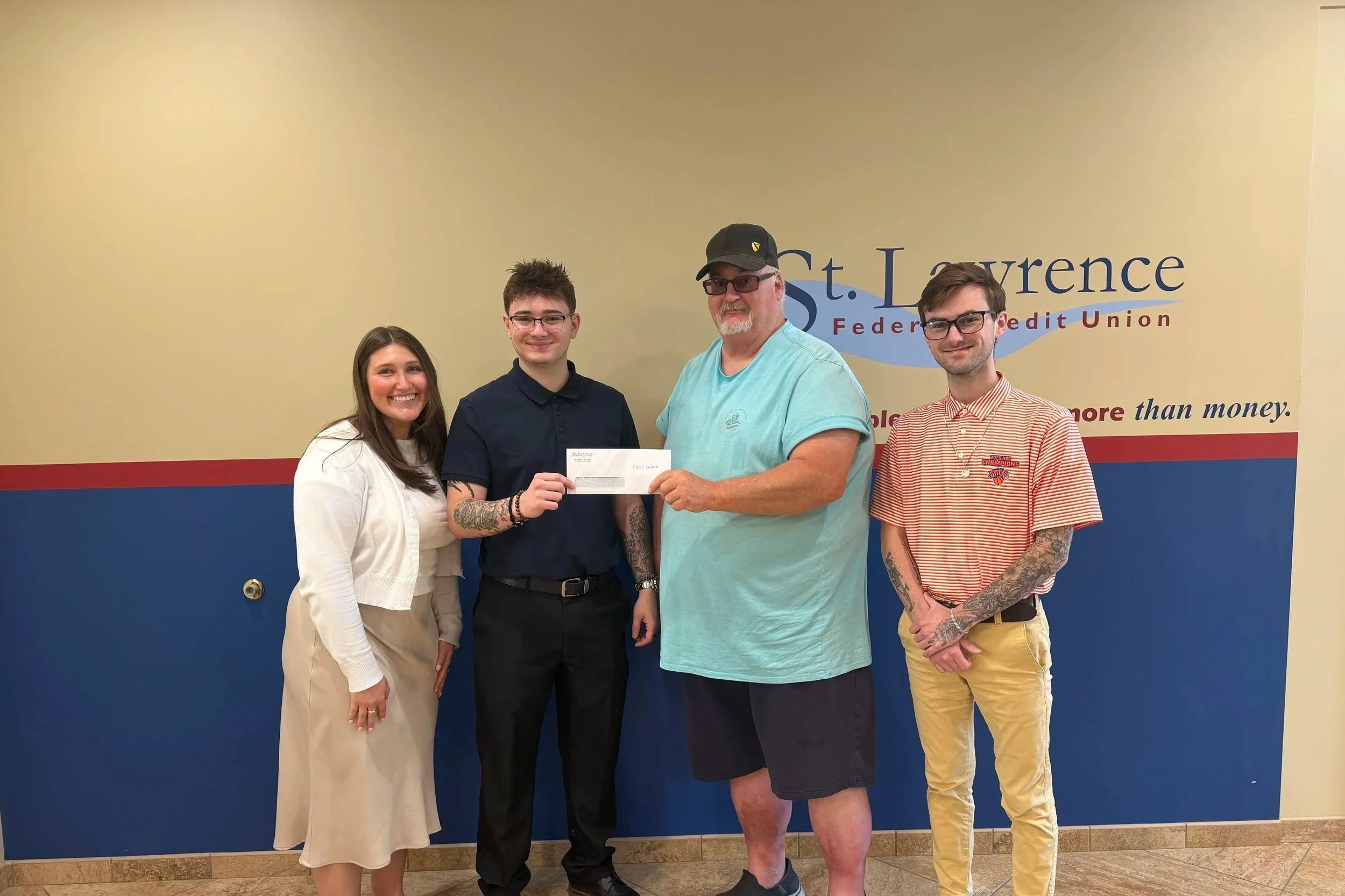 A young adult female, and two young adult males presenting a donation check to an adult male for a community event.