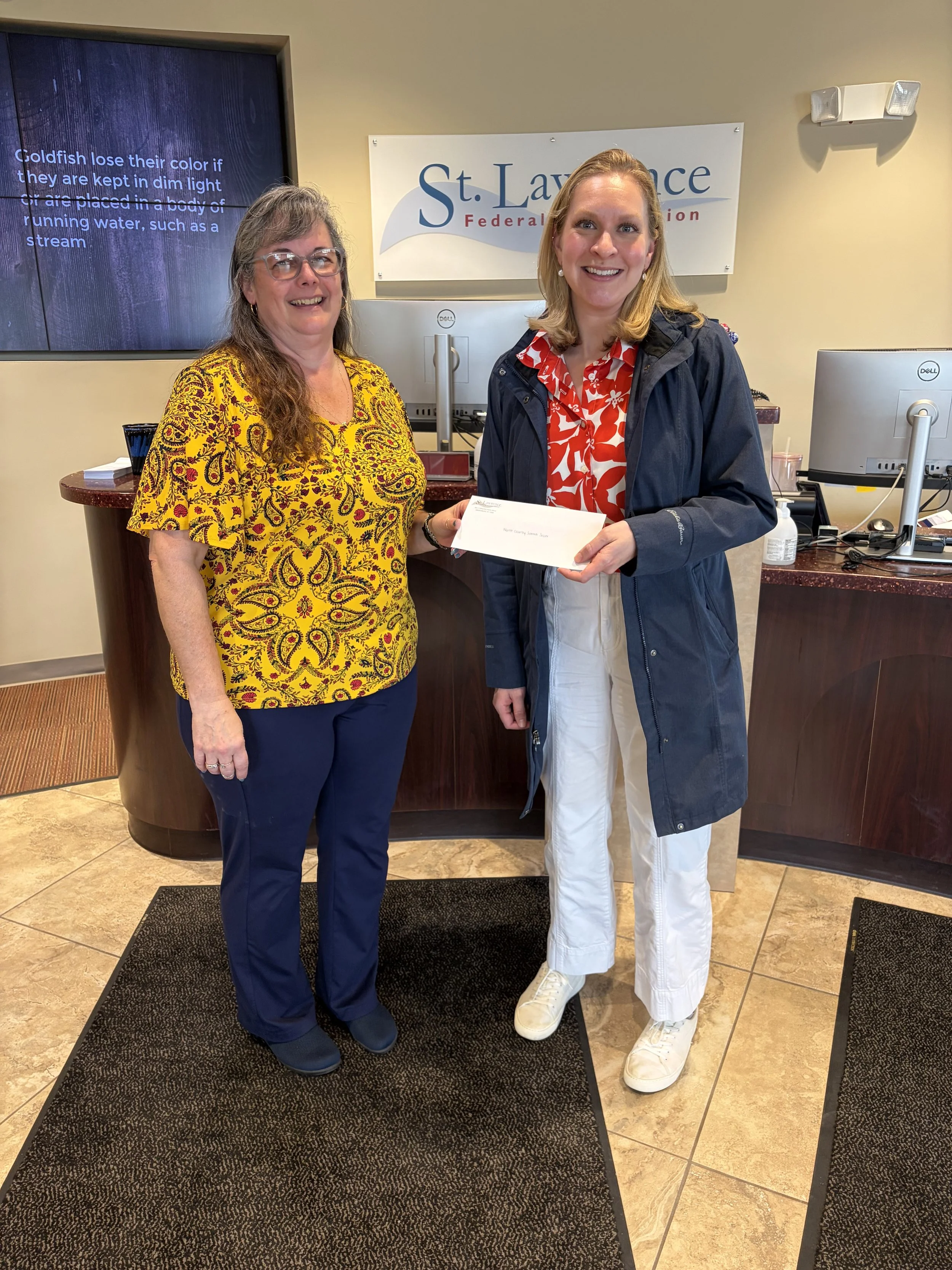 One adult female accepting a donation check from another adult female in support of the Women's Summer Soccer League in the North Country.
