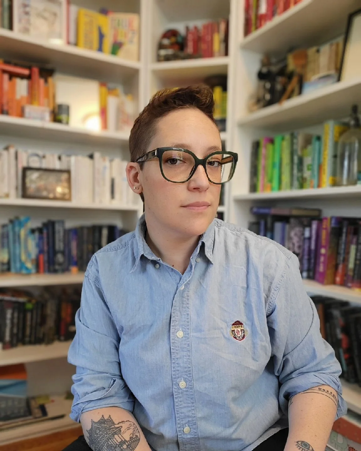 Sara has light skin, brown hair and is wearing green glasses and a denim shirt with the sleeves rolled up, revealing black and white tattoos. Behind them is a colorful bookshelf.