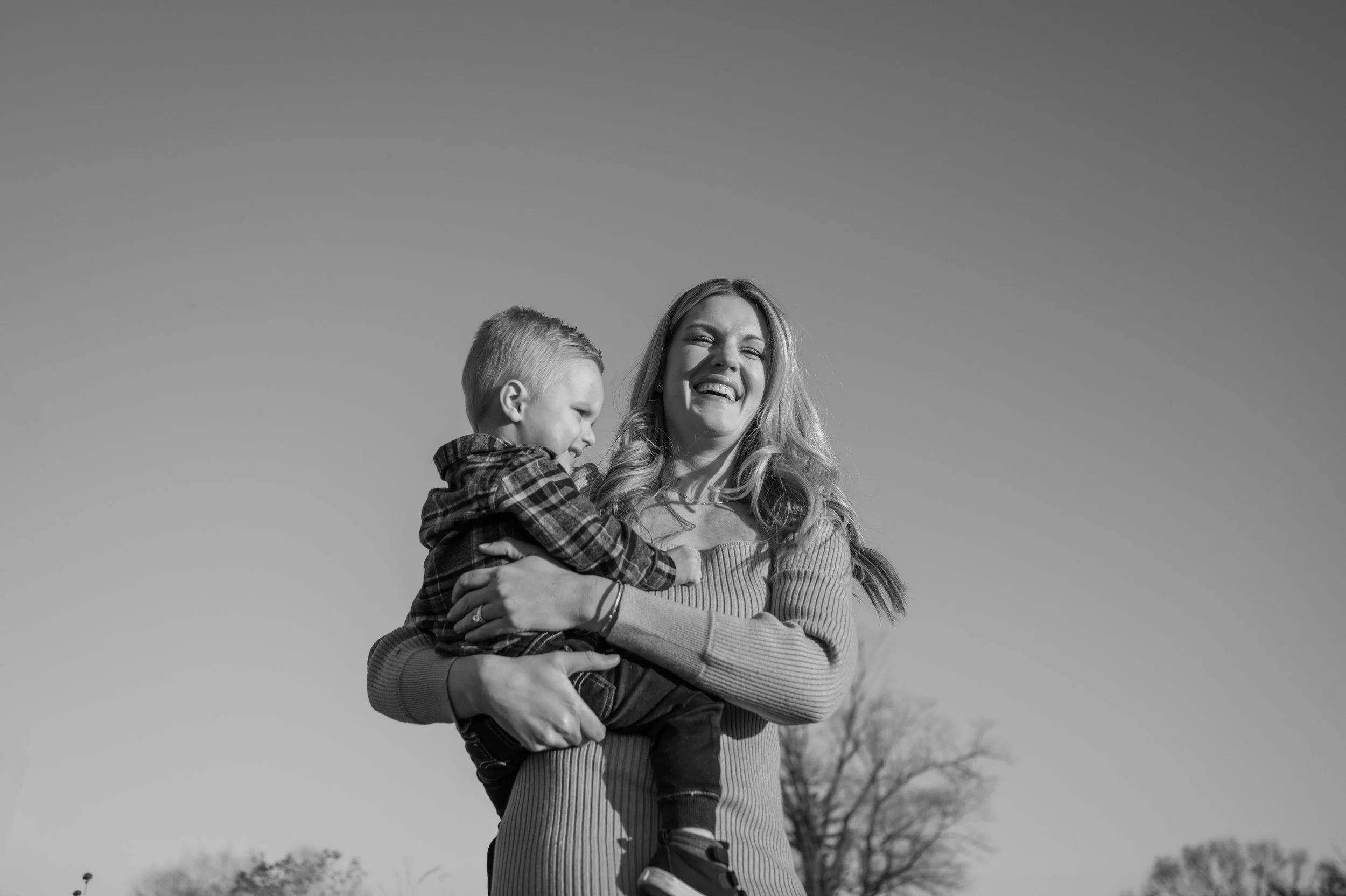 black & white image of mother laughing while holding her toddler son outside with the sun on their faces and clear sky behind them.