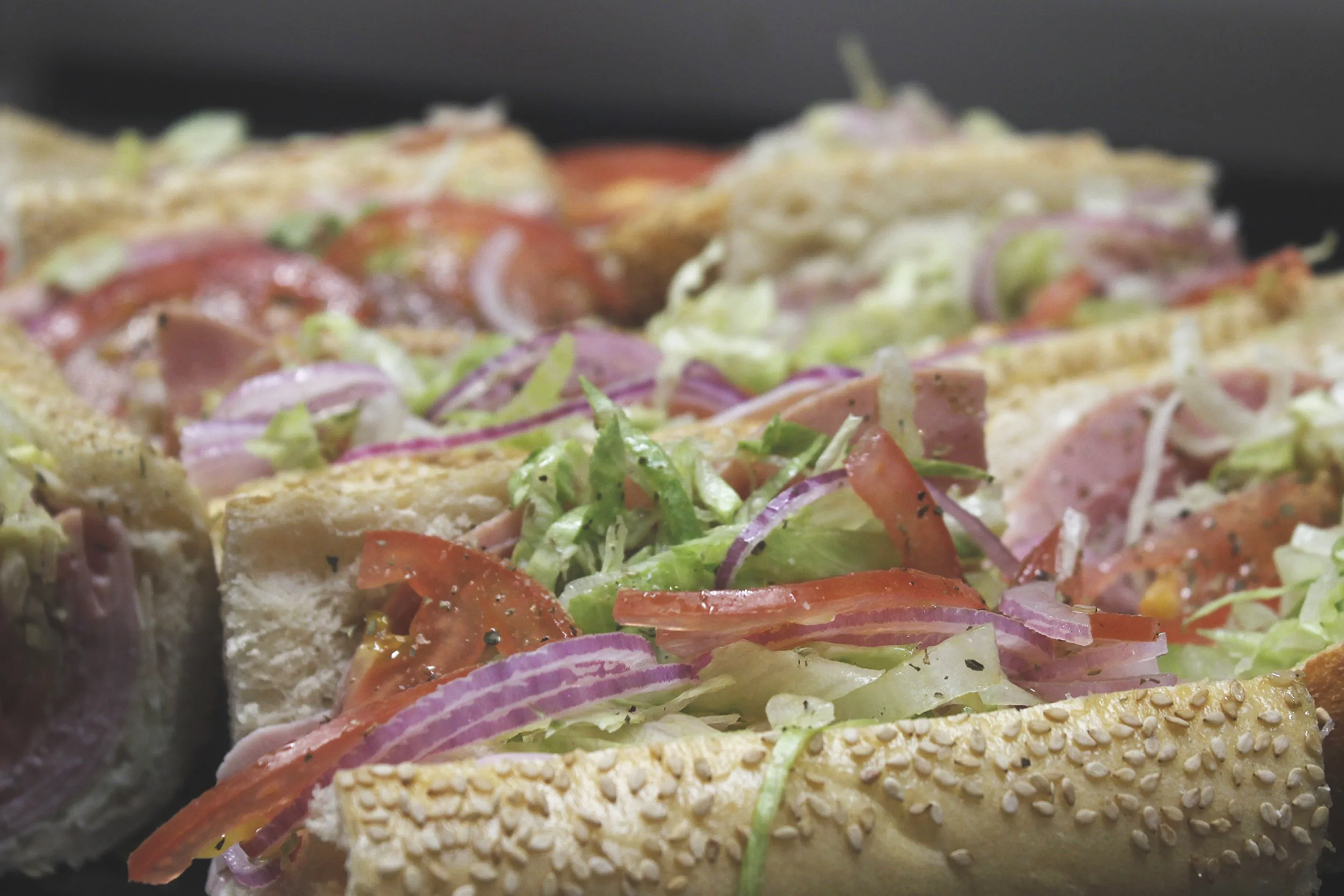 Close-up of a sandwich with sesame seed bread, filled with ham, lettuce, tomato, and red onion.