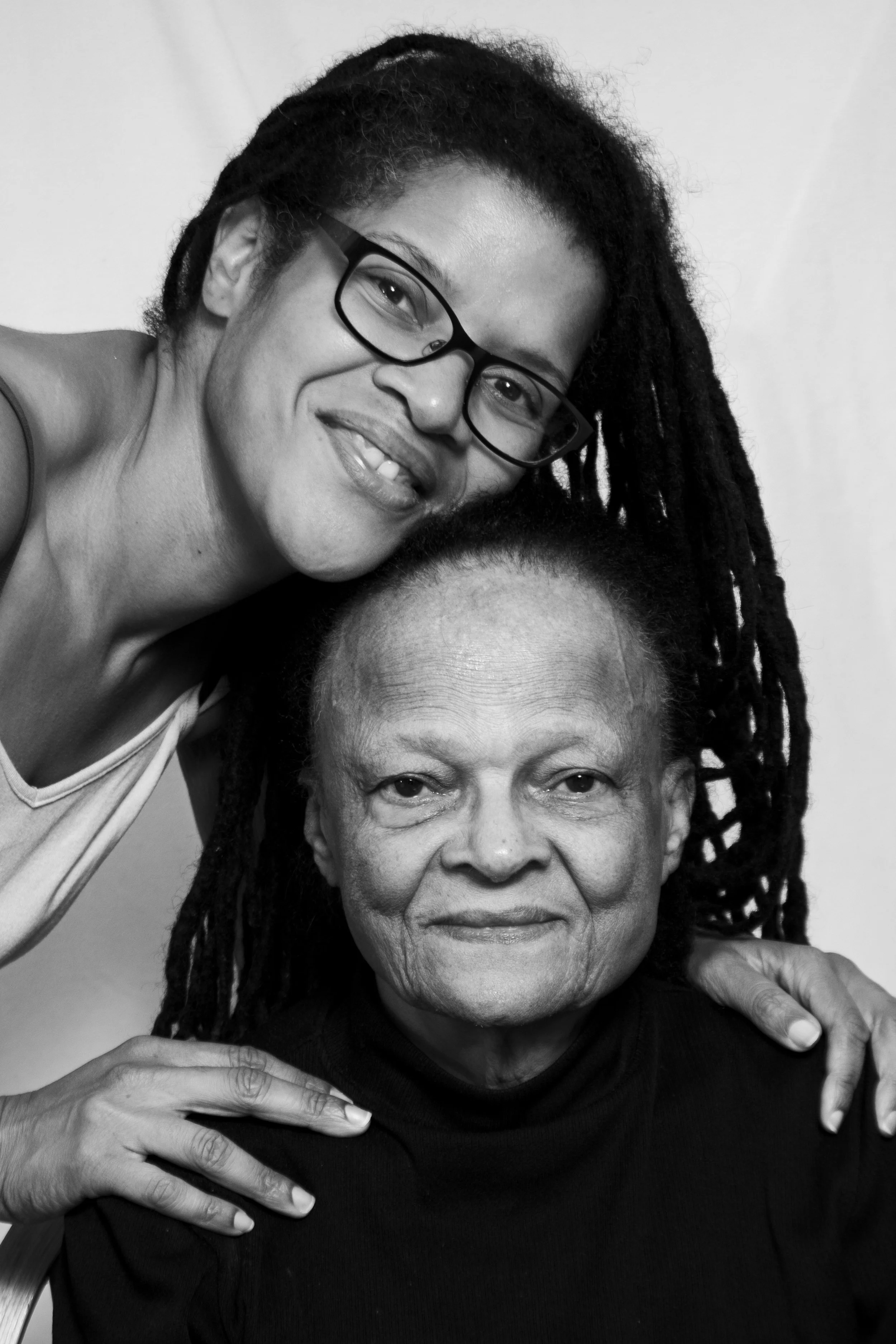 A black and white photo of two women, one younger with glasses and dreadlocks smiling, and an older woman with short hair and a calm expression, both closely positioned and looking at the camera.