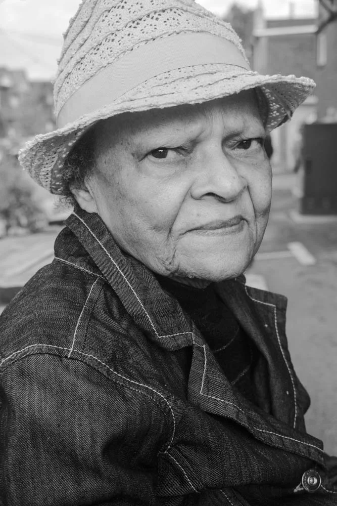 Close-up black and white photo of an elderly woman wearing a straw hat and a denim jacket, sitting outdoors.