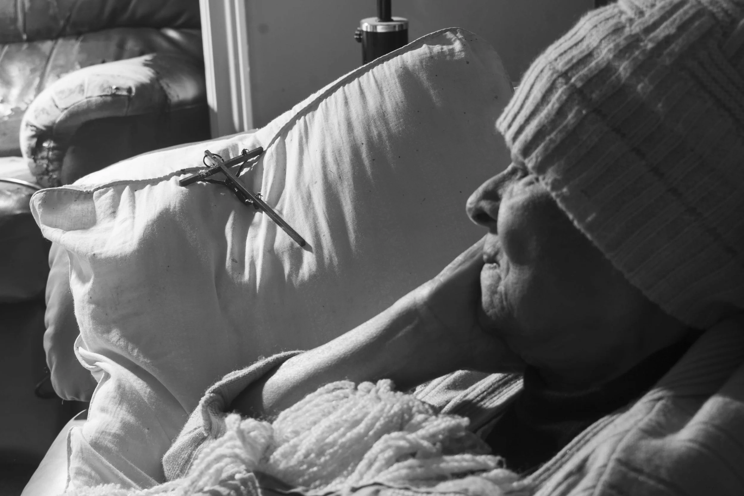 A person dressed in warm clothing, including a striped knit hat, is sleeping on a couch covered with a blanket. There are pillows and a crucifix resting on the person's chest.