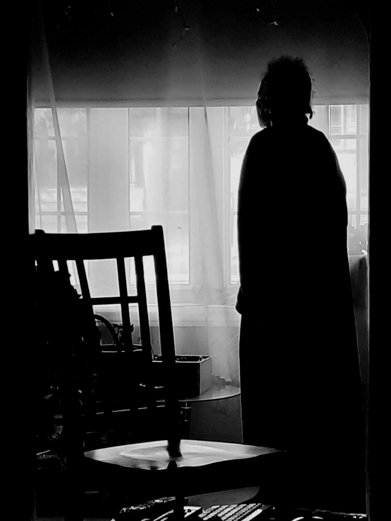 A person standing in front of a large window with curtains, the back facing the camera, in silhouette. The room appears to have chairs and possibly a table, all in shadow, with natural light coming through the window.