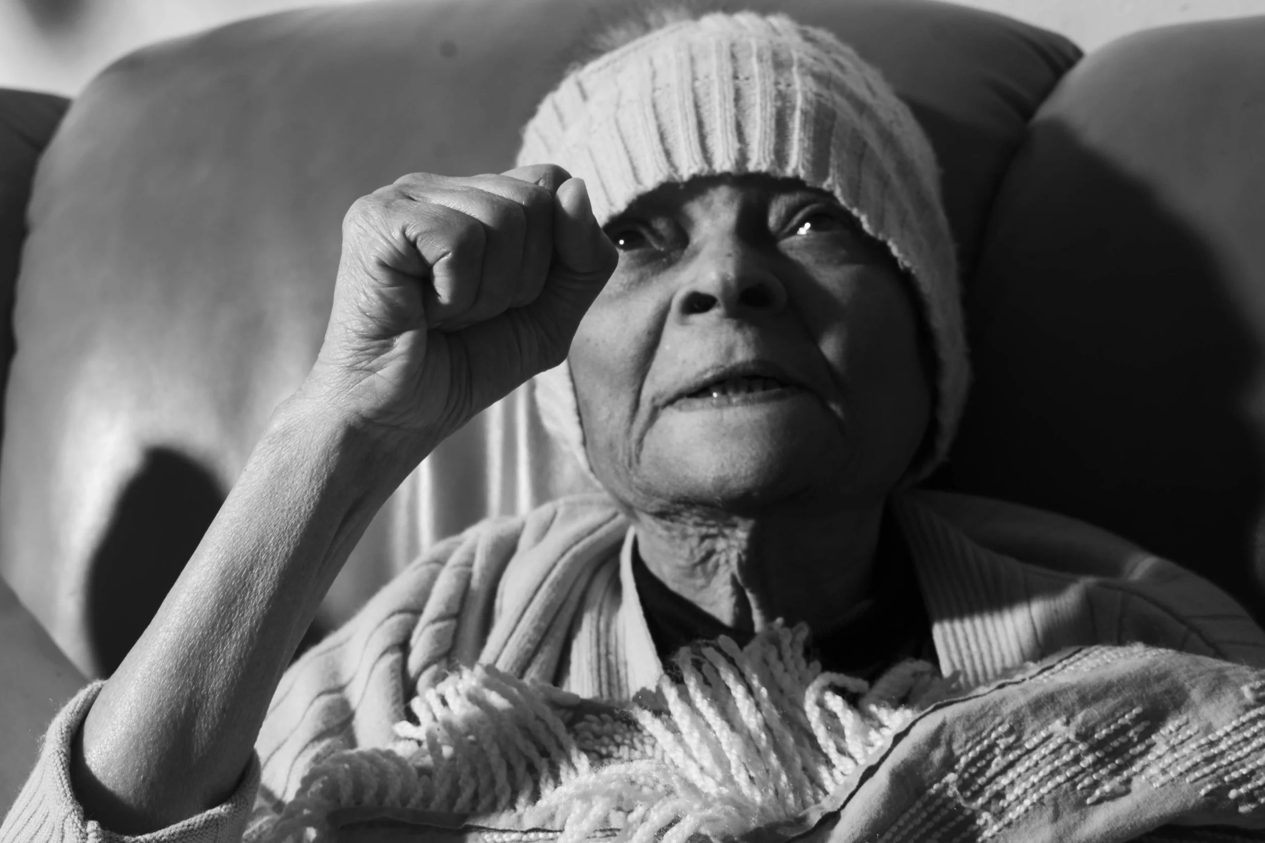 An elderly woman lying down wearing a knit hat and sweater, looking up with her hand near her eye in a black-and-white photograph.