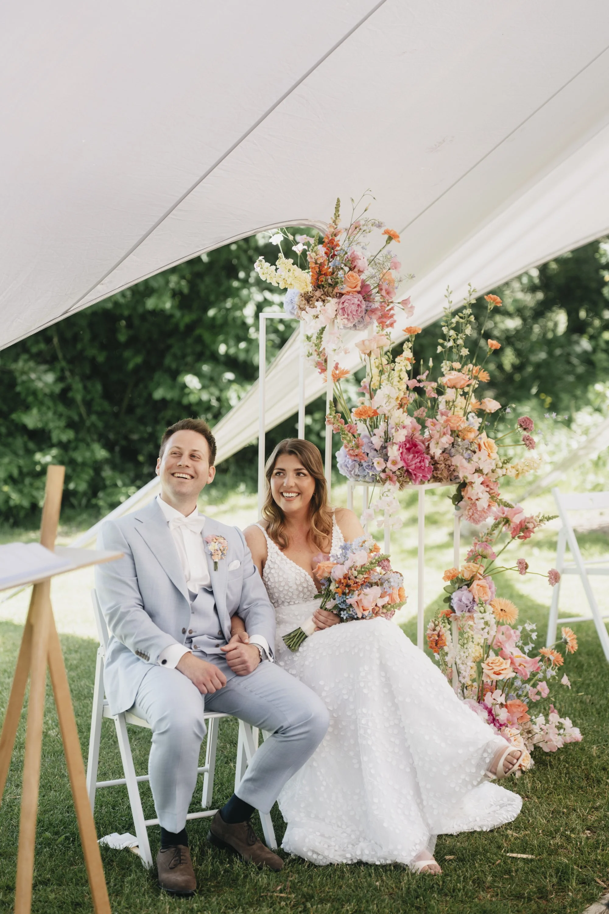 Hochzeitsfotograf Münster | Paul Popanda Hochzeitsreportage Paderborn. Glückliches Brautpaar sitzt bei einer sommerlichen Hochzeitsfeier unter einem weißen Zelt, umgeben von farbenfrohen Blumenarrangements.