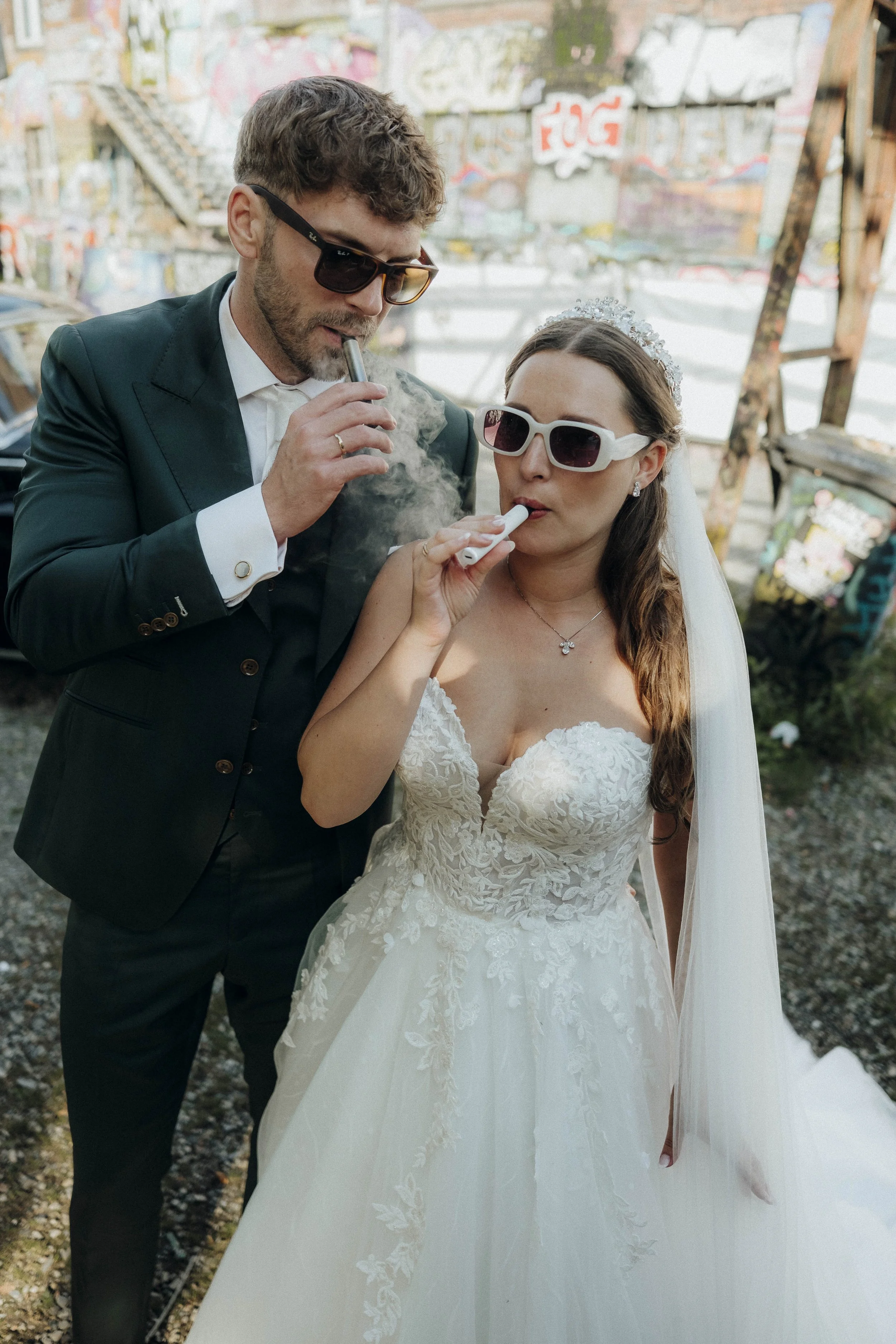 Brautpaar mit Sonnenbrillen raucht gemeinsam auf einer Hochzeit im Freien.