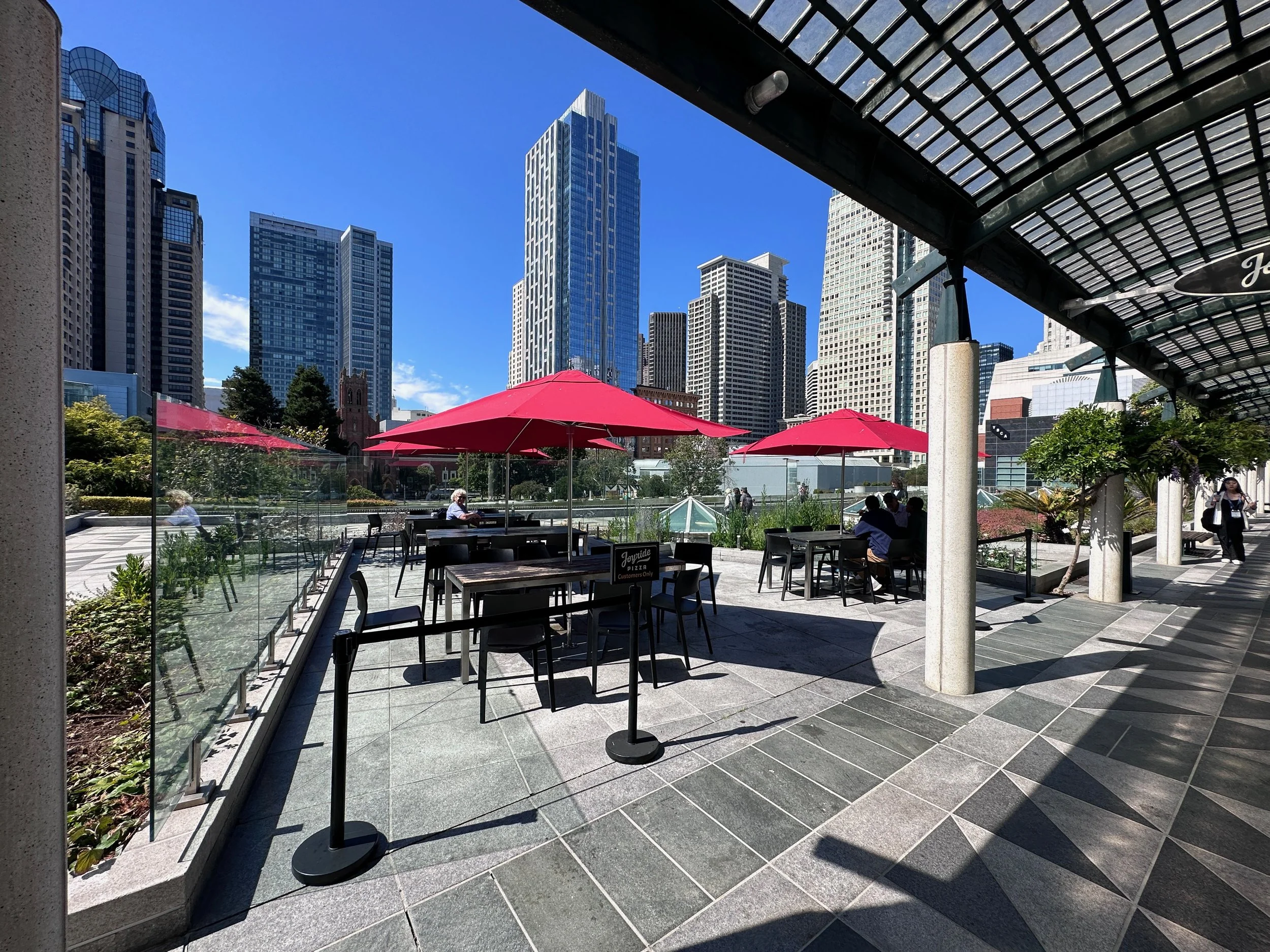 Outdoor seating area with tables, chairs, and red umbrellas in a cityscape with tall skyscrapers and people sitting and walking under a glass roof.