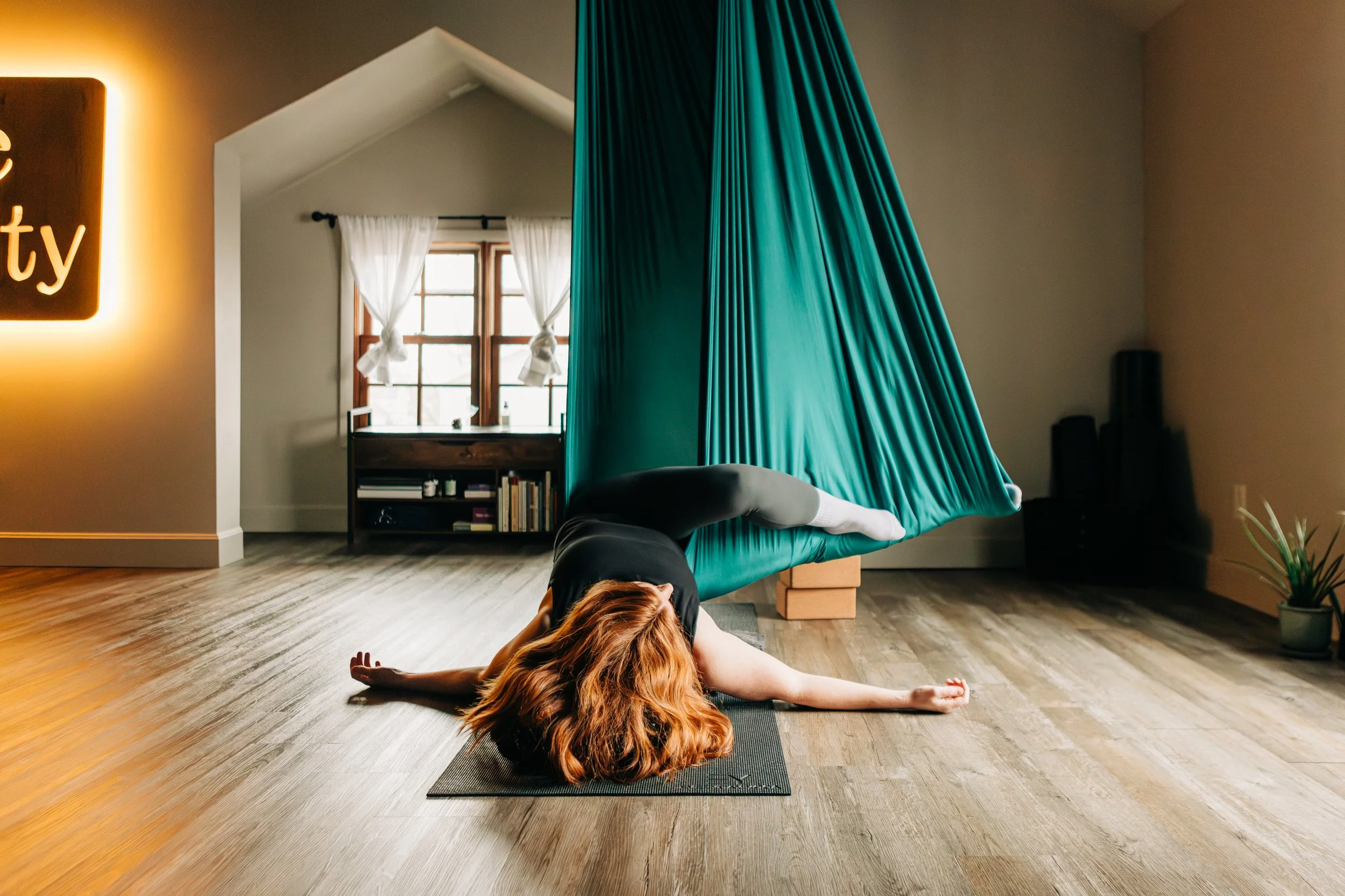A woman practicing yoga in a room with wooden floors and large windows, engaging in a challenging pose using a teal hanging fabric hammock.