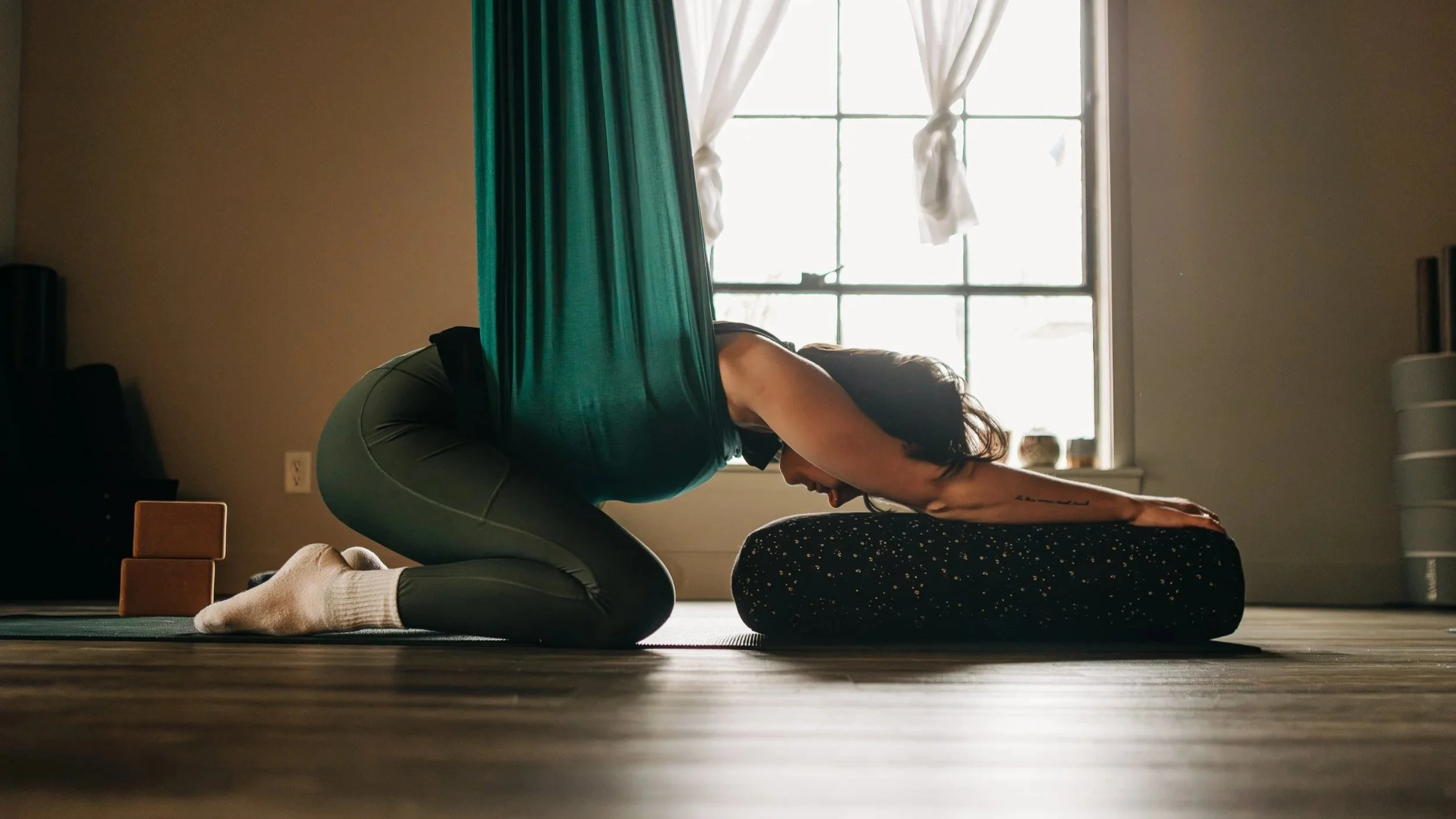 A woman practicing yoga on a mat in a room with sunlight coming through a window, using a bolster for support.